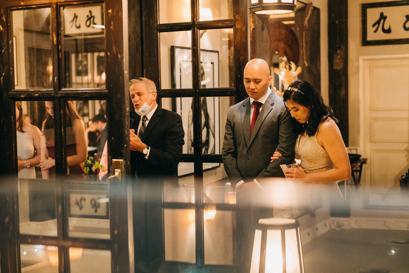 A man in a suit speaks at a wedding reception at The English House Singapore, while a couple stands beside him, looking down. The elegant setting features warm lighting and reflections.