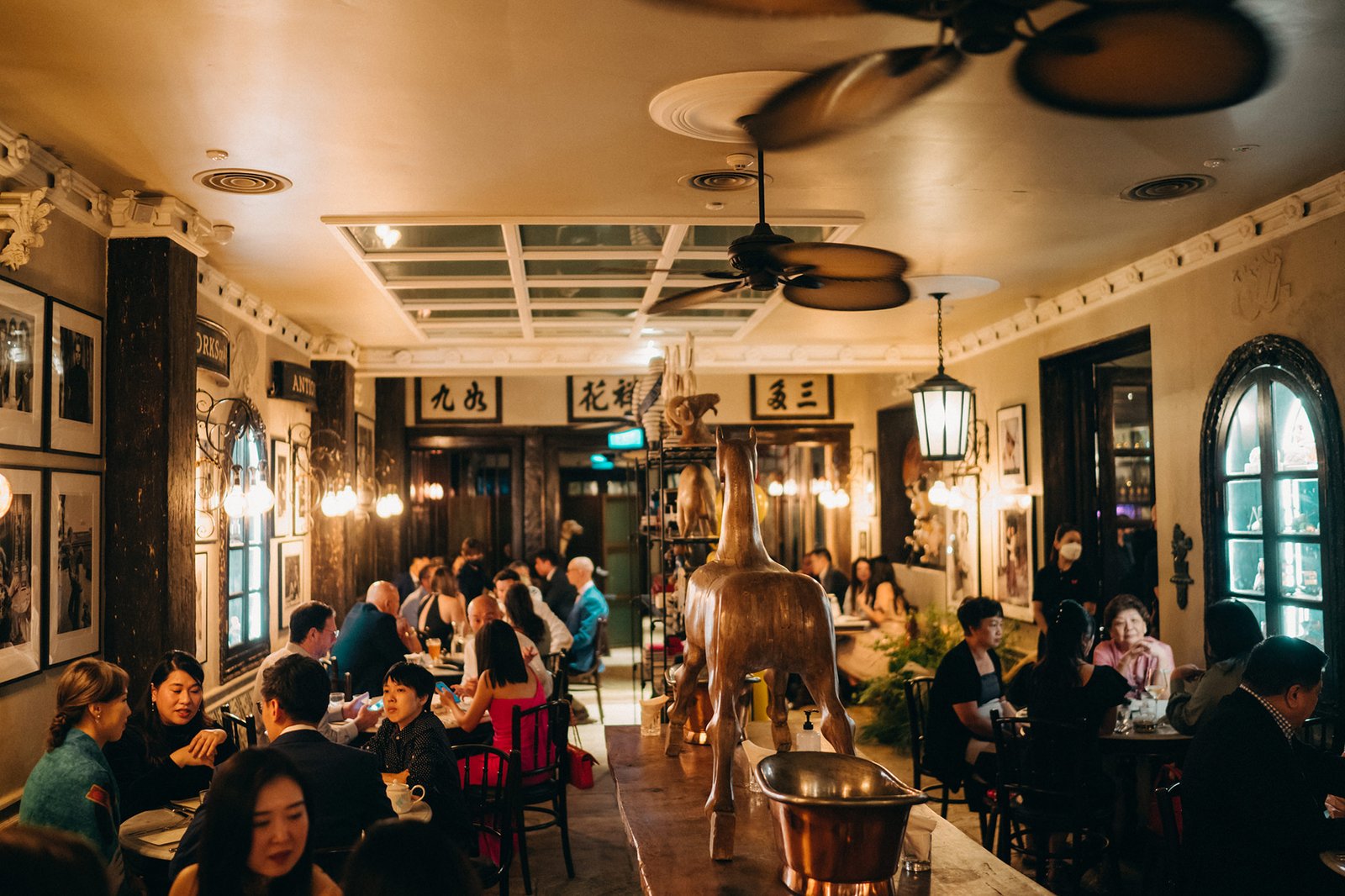 A warm, dimly lit interior of The English House, a restaurant in Singapore, with patrons dining at tables adorned with copper accents. Elegant decor, including ornate lamps and a large horse sculpture, creates a sophisticated ambiance.