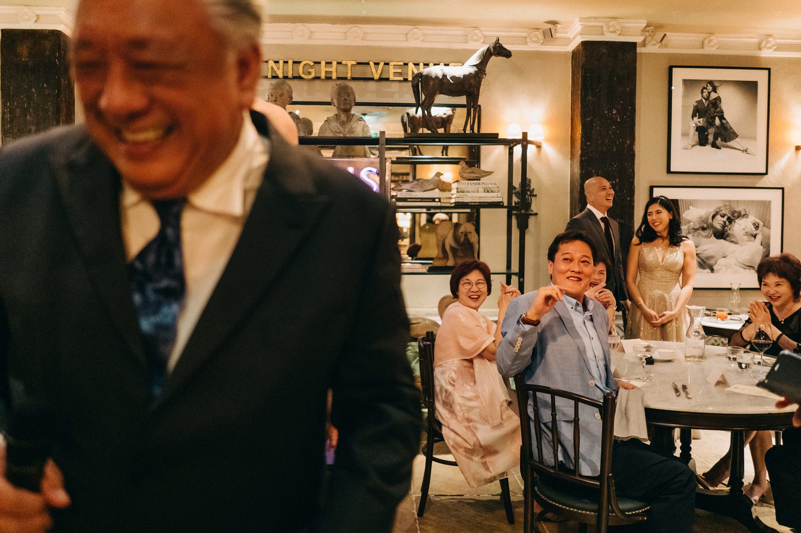 A joyous wedding reception at The English House Wedding Singapore, with guests laughing and celebrating around elegantly set tables. A distinguished man in a dark suit smiles broadly in the foreground, with other attendees in formal attire visible in the warm, inviting ambiance.