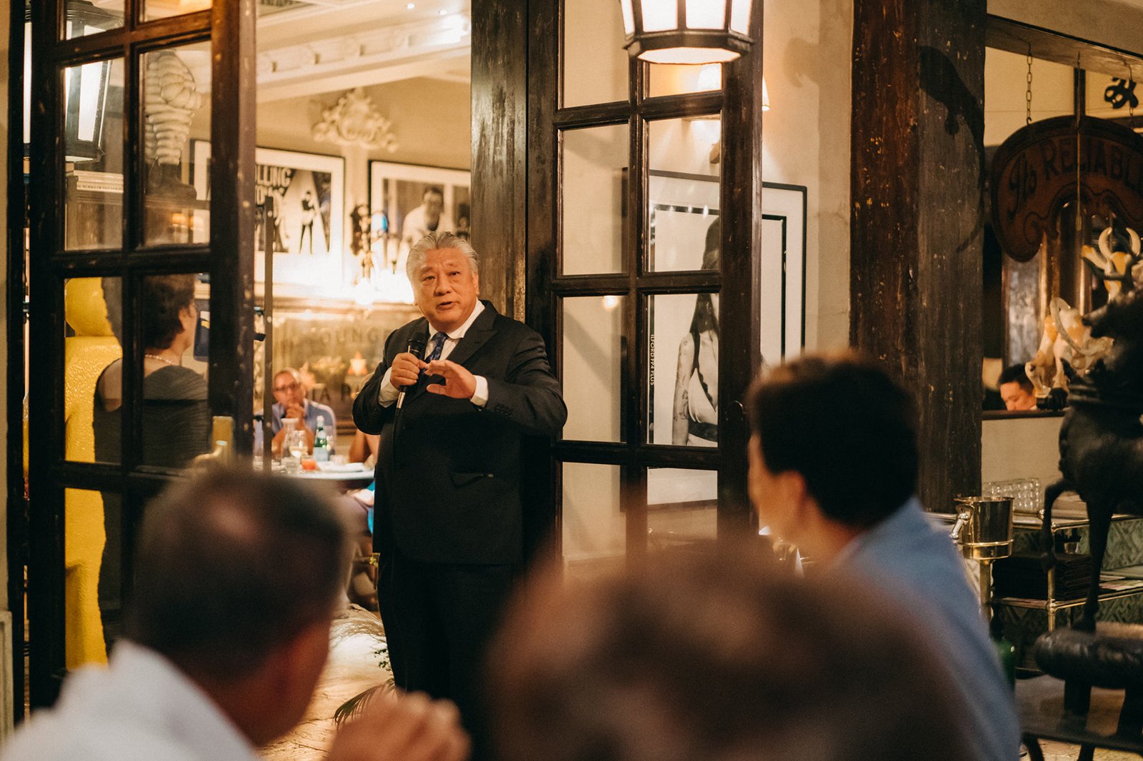 A distinguished Asian man in a suit gives a speech at an event, holding a microphone. The background shows a warmly lit interior with large windows and framed artwork, hinting at a sophisticated gathering like The English House Wedding Singapore.
