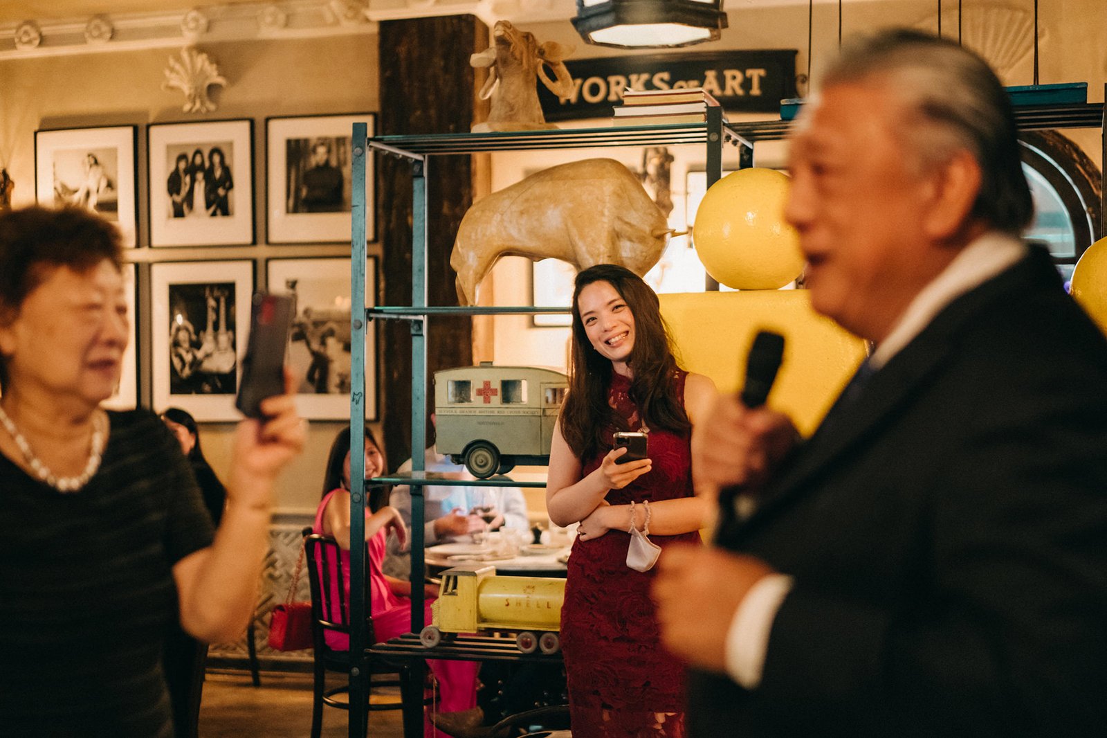 A smiling young woman in a red lace dress holds a phone at The English House Wedding Singapore, with guests and artwork in the background.