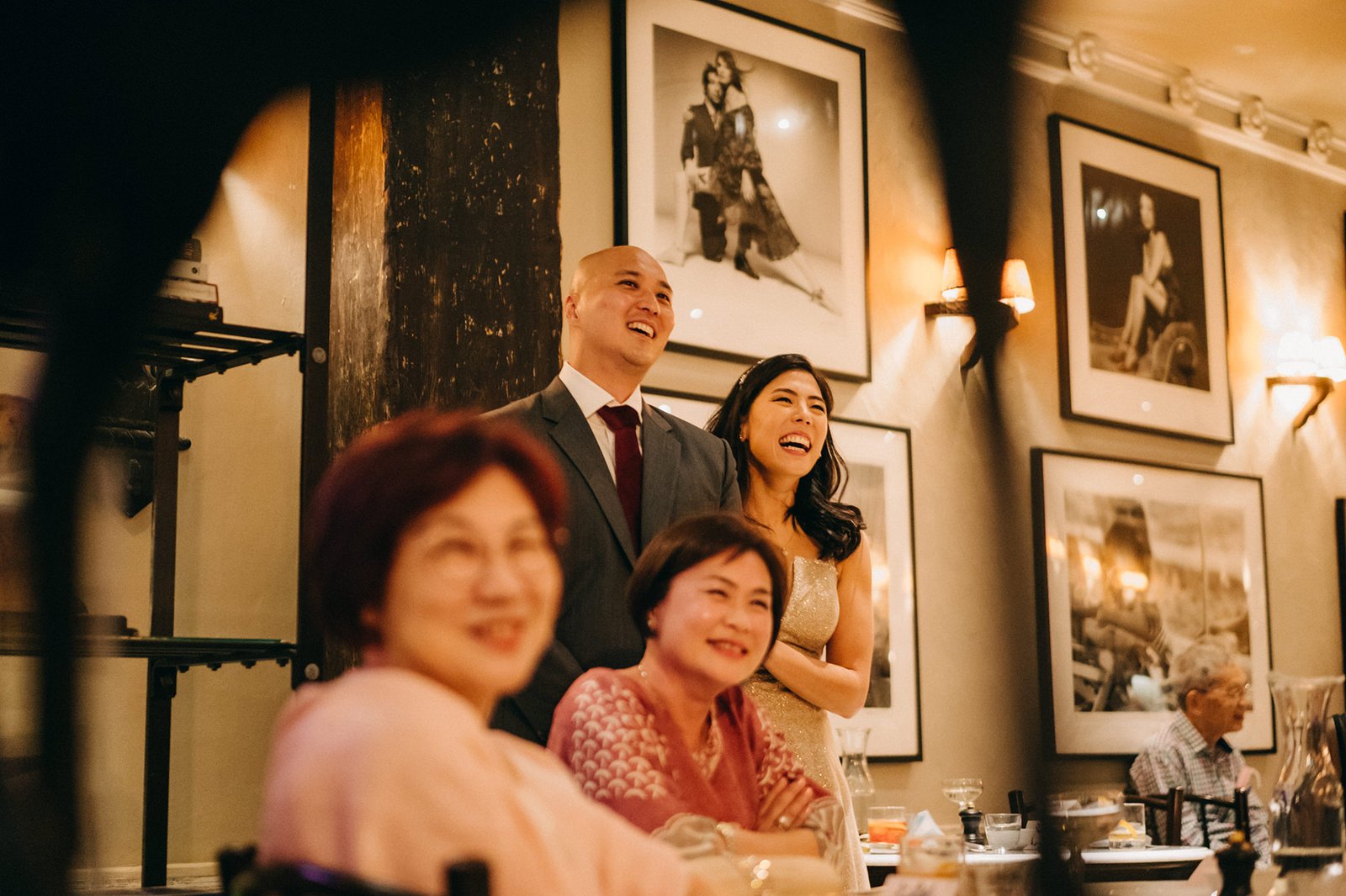 A joyful couple, Kin and Christine, laugh during their wedding reception at The English House, Singapore, with guests in the foreground.