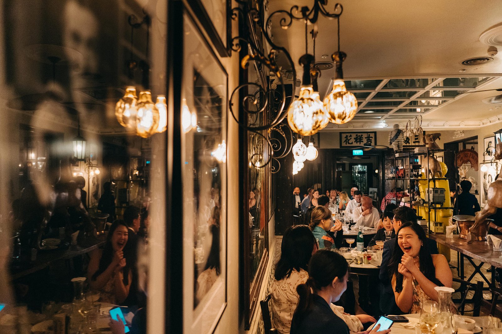 A lively wedding reception at The English House Singapore, where guests laugh and enjoy a meal, with warm lighting from ornate chandeliers reflecting on the walls.