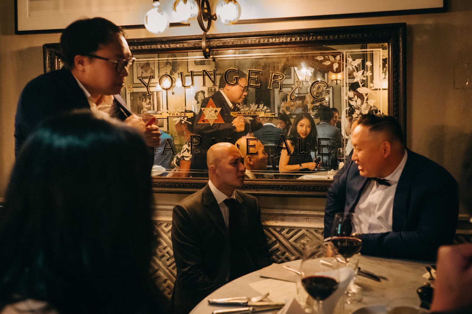 Guests in formal attire converse at a wedding reception at The English House Singapore, reflected in a mirror with "YOUNGER & CO TRADE MARK" visible.