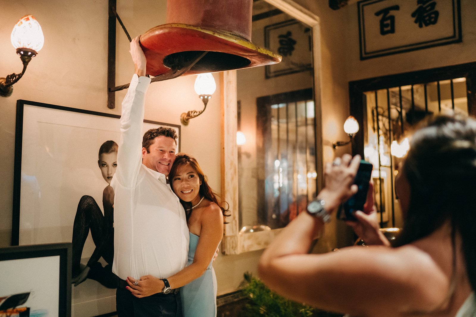 Couple posing under a large red hat at The English House Wedding Singapore, with a woman taking a photo on her phone.
