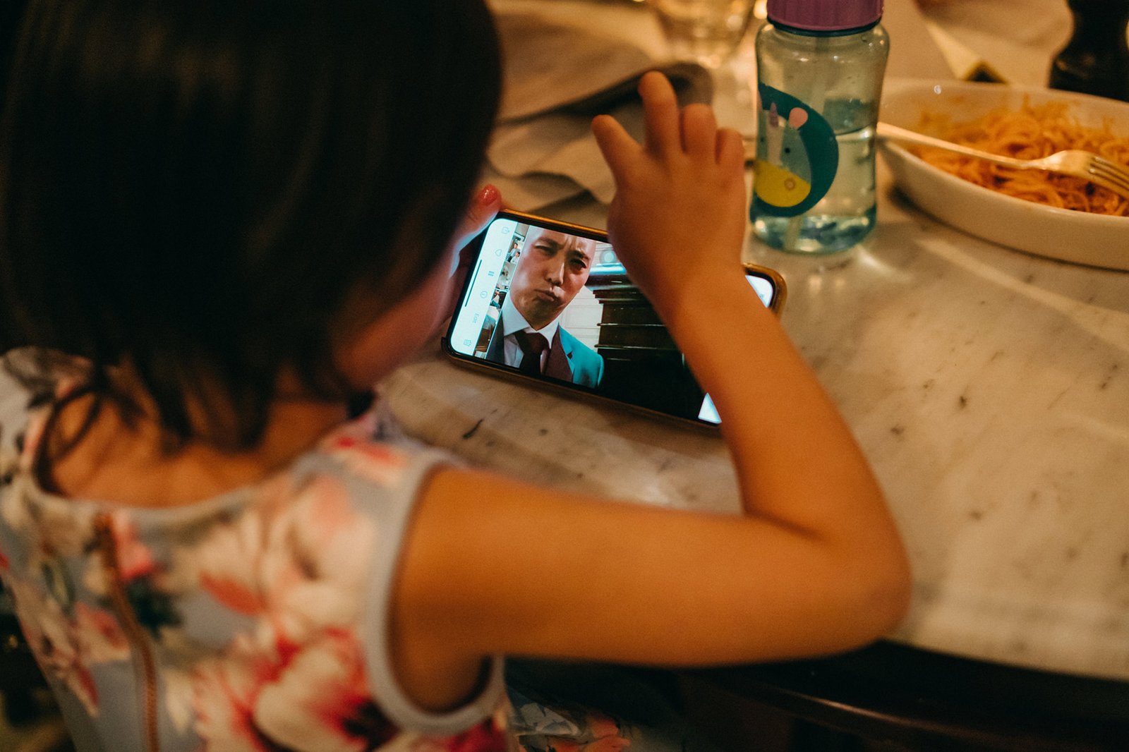 A young girl in a floral dress watches a video on a smartphone at a wedding reception table, with a bottle of water and a plate of cacio e pepe pasta in the background. The English House Wedding Singapore.