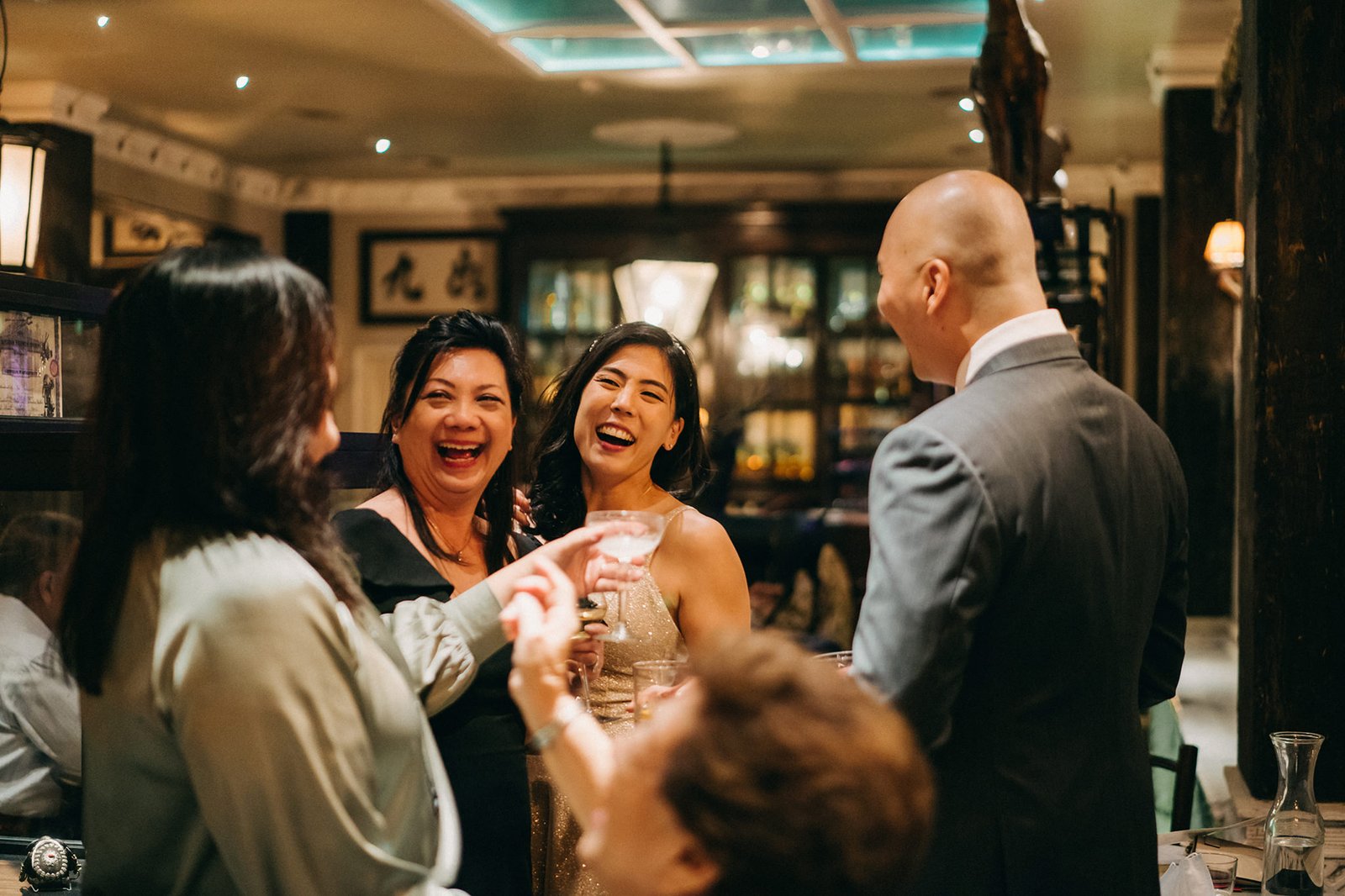 A joyful wedding reception at The English House Wedding Singapore, where a bald man in a suit toasts with guests, capturing a moment of celebration and laughter among friends.