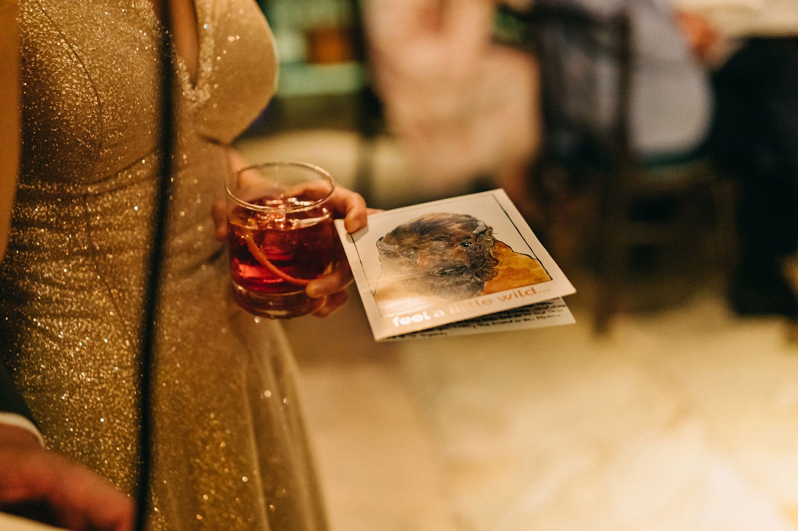 A guest at The English House wedding in Singapore holds a drink and a card with "feel a little wild..." text, wearing a sparkly gold dress.