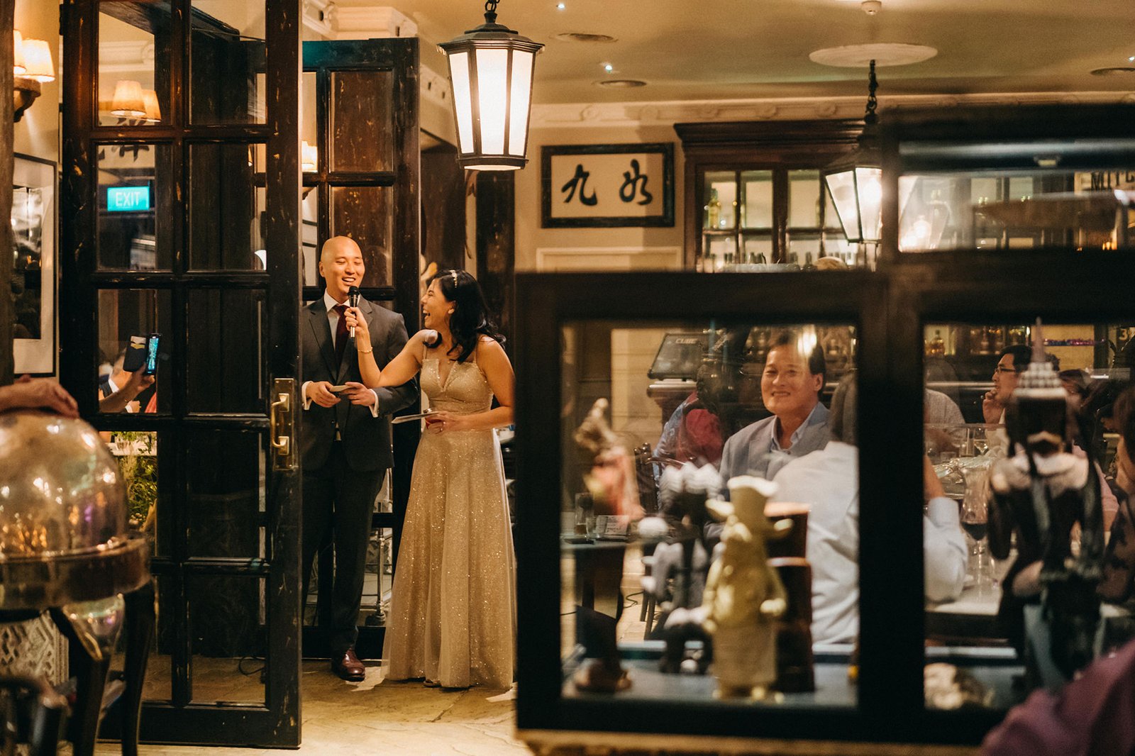 A smiling man in a suit and a woman in a sparkling gown speak at a wedding reception at The English House Wedding Singapore, with guests in the background.