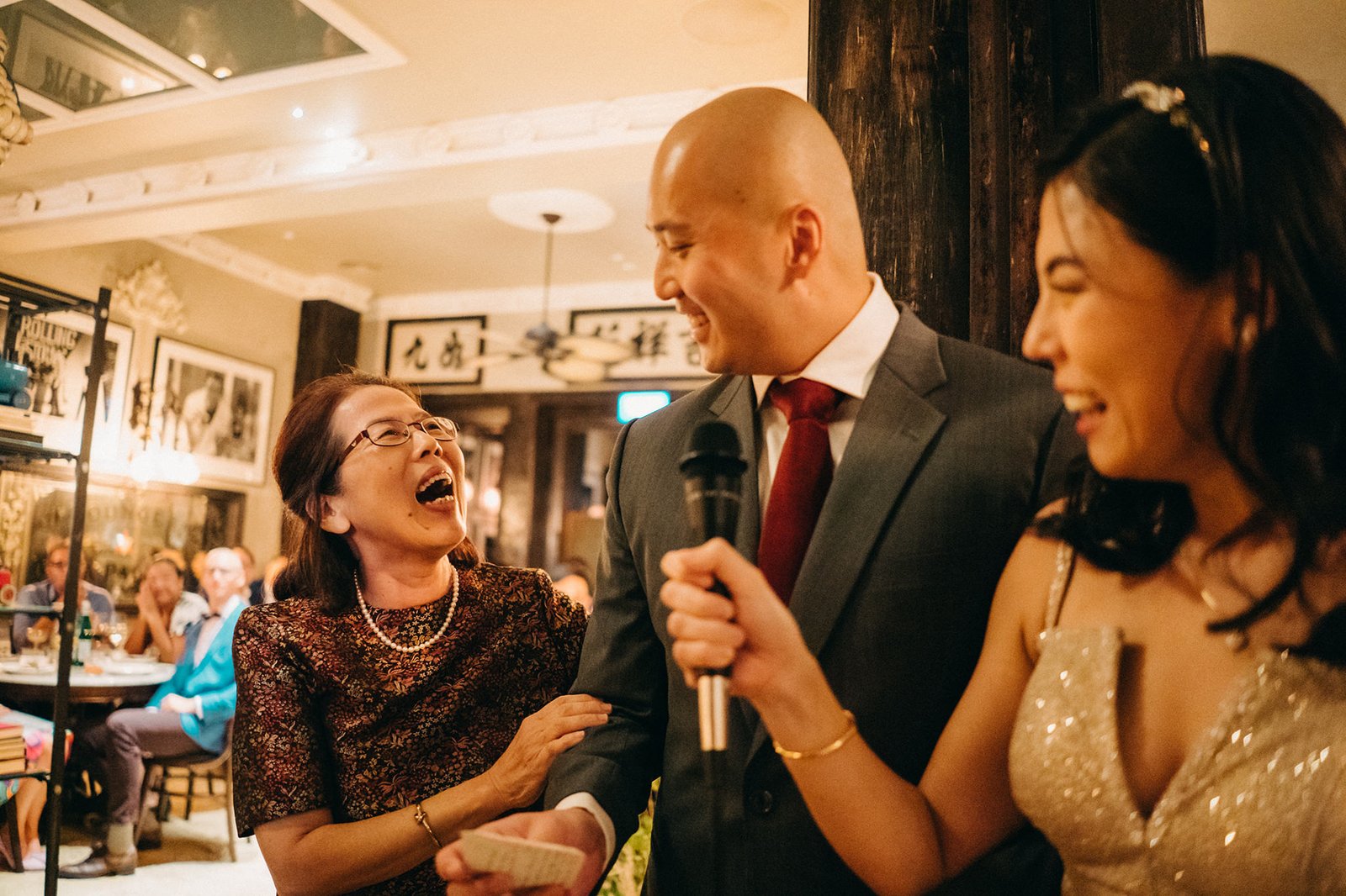 A joyous wedding reception at The English House Wedding Singapore, with a man in a suit speaking into a microphone, flanked by two laughing women, one in a sparkly dress.