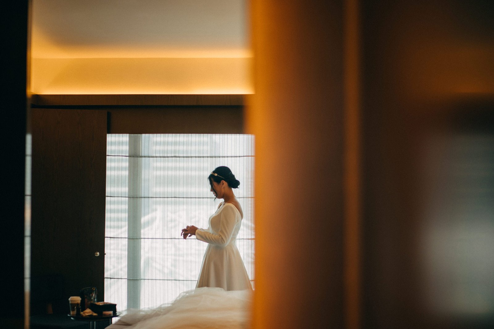 A bride in a white wedding dress with long sleeves and a tiara stands near a window, preparing for her Keyaki Garden Pavilion Wedding in Singapore.