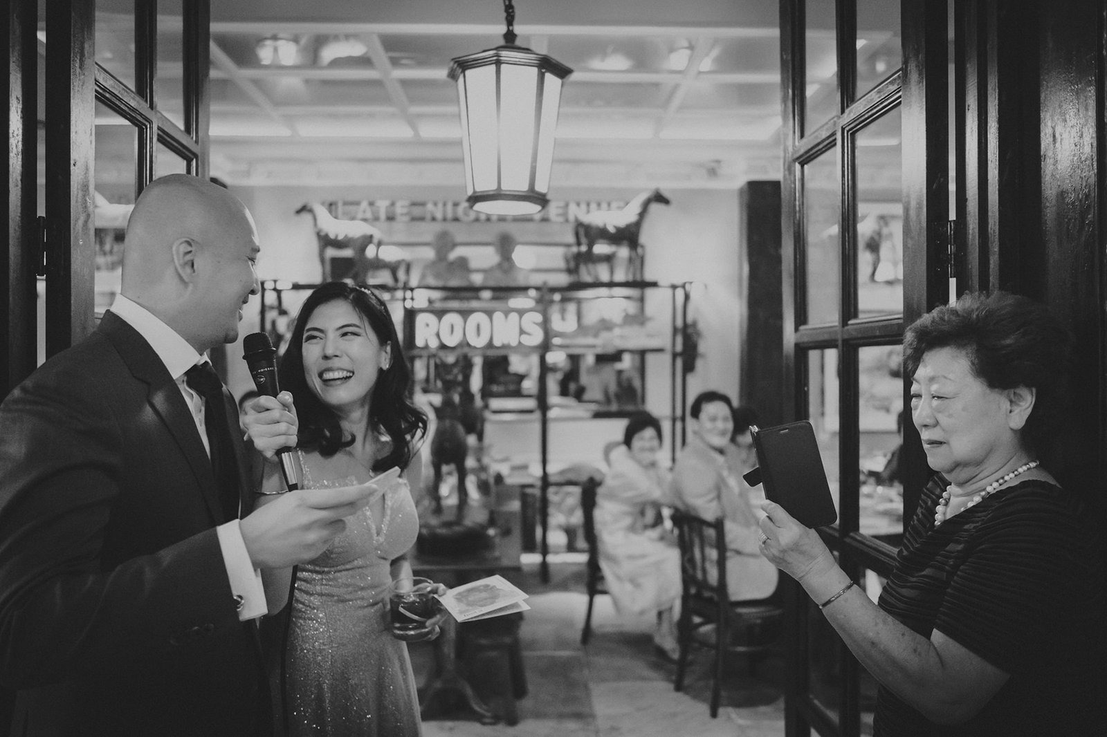 A wedding MC speaks into a microphone at The English House Wedding Singapore, while a bride laughs and an older woman holds a tablet, at a celebration with a vintage feel.