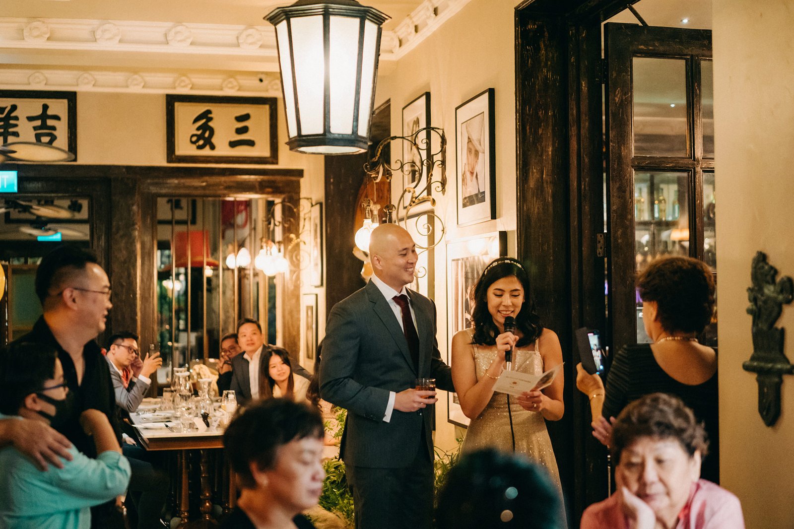 A bride in a sparkling gold dress gives a speech with her groom in a suit beside her at The English House Wedding Singapore. Guests watch and listen in the elegant, dimly lit reception hall.