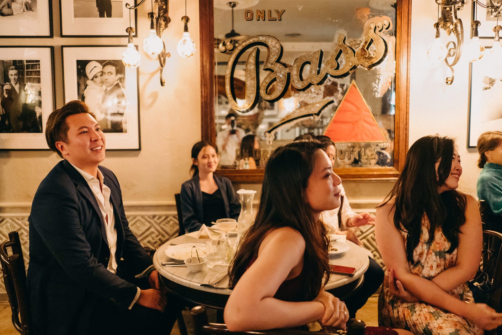 A celebratory gathering at The English House Singapore, with guests enjoying a meal. A man in a suit smiles, while women in dresses are seated at tables, with vintage decor and a "Bass Bottle" sign in the background.