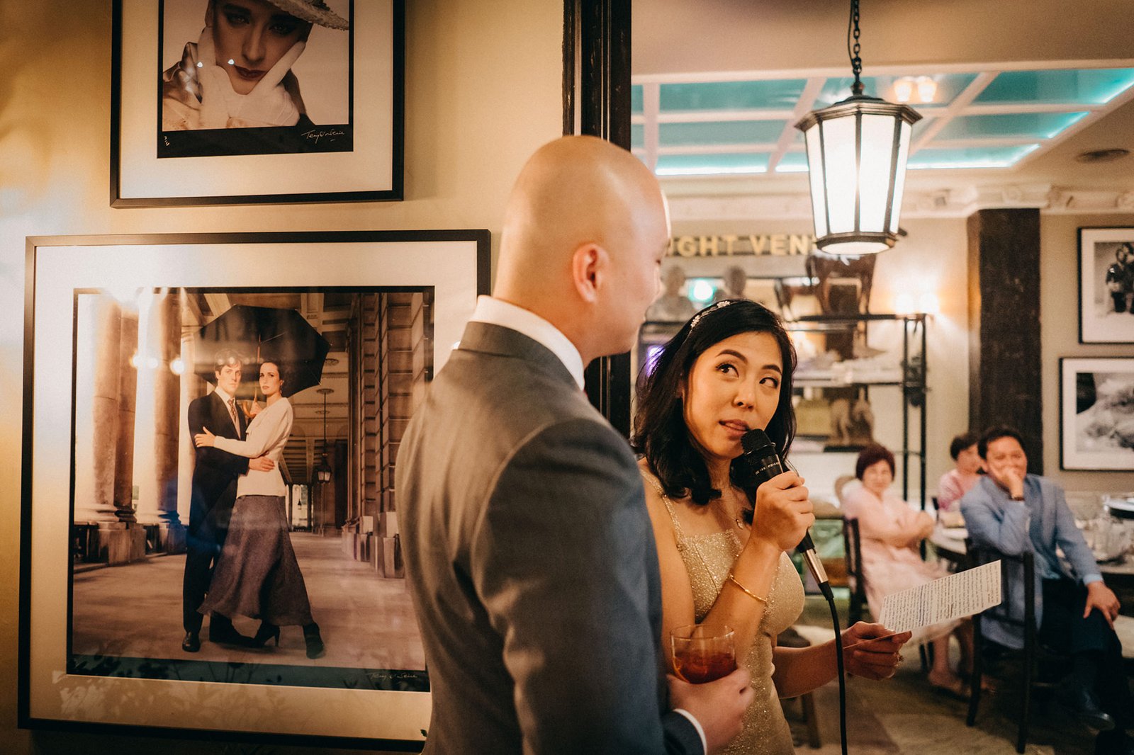 A woman in a sparkling gold dress speaks into a microphone at The English House Wedding Singapore, holding a drink and a paper. A man in a grey suit stands beside her, facing away from the camera.