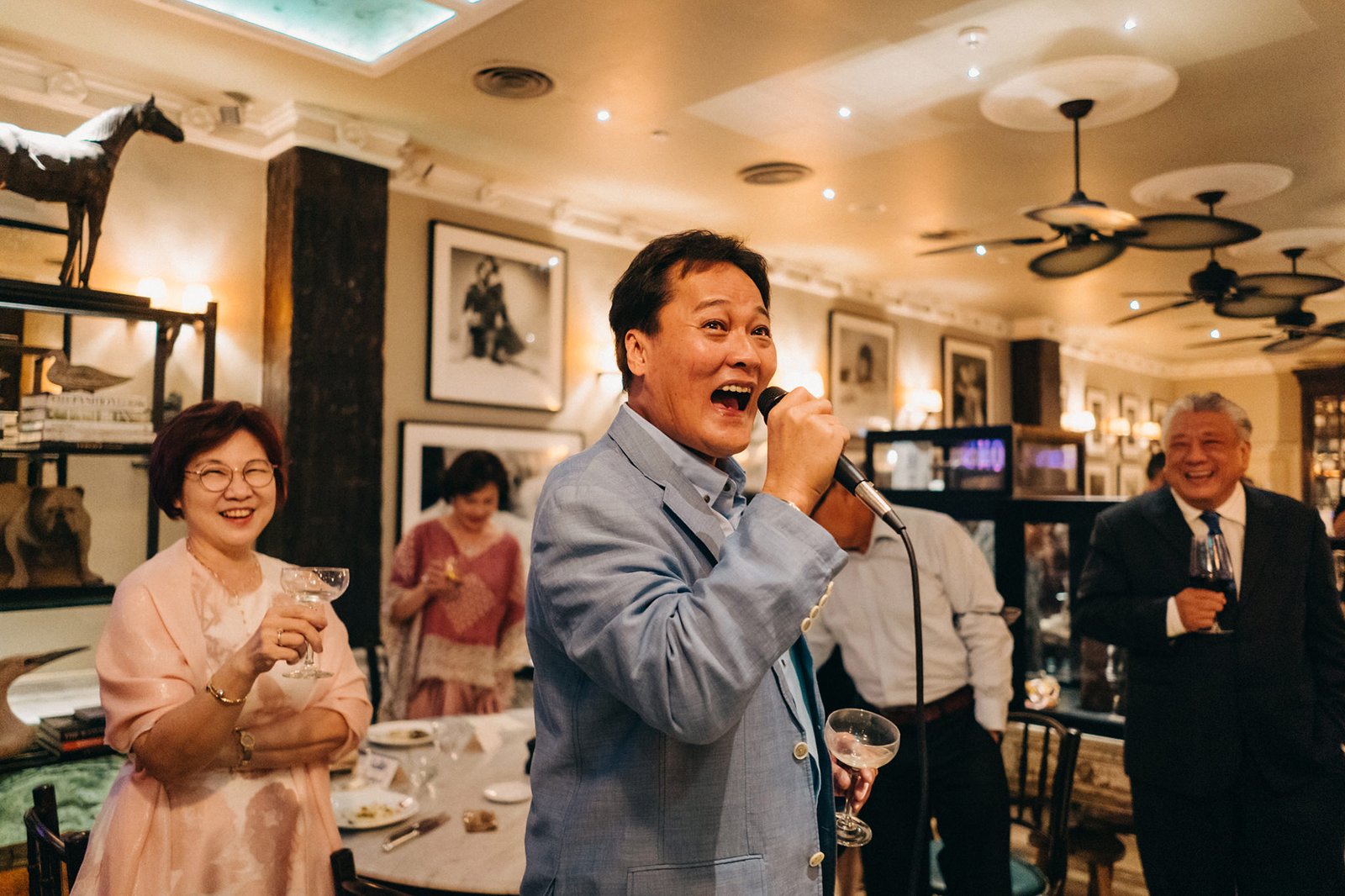 A man in a light blue suit speaks into a microphone at a wedding reception at The English House, Singapore, with other guests smiling in the background.