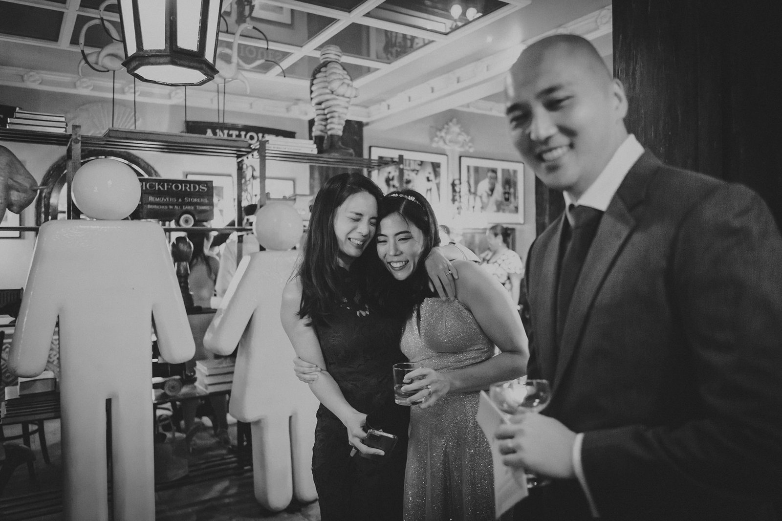 Black and white photo of a happy Asian couple embracing at The English House Wedding Singapore, with a man in a suit smiling in the foreground.