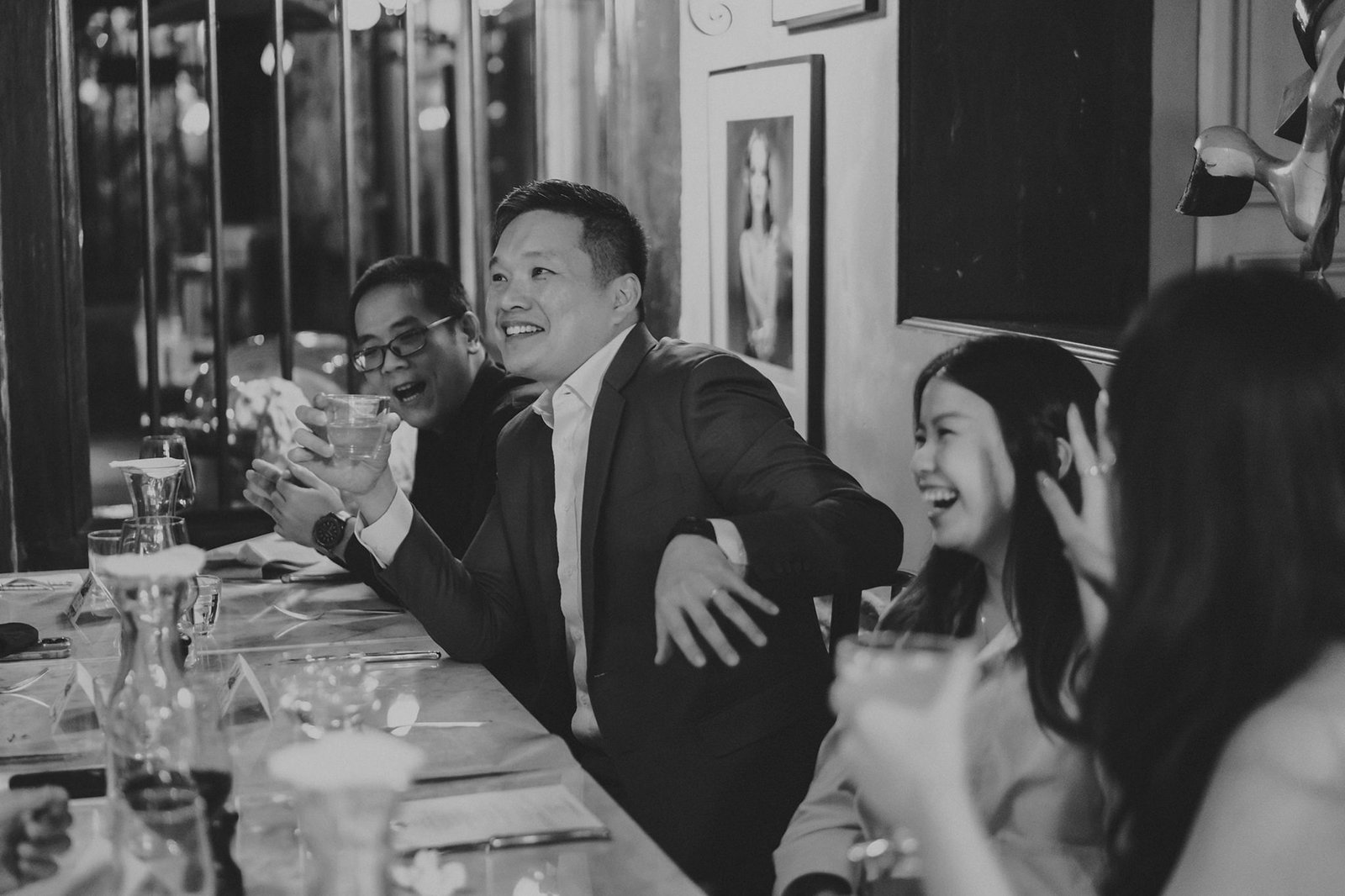 A lively group of friends and family laugh and toast at a celebratory dinner, possibly a wedding reception, at The English House Wedding Singapore. The black and white photo captures a candid moment of joy and connection.