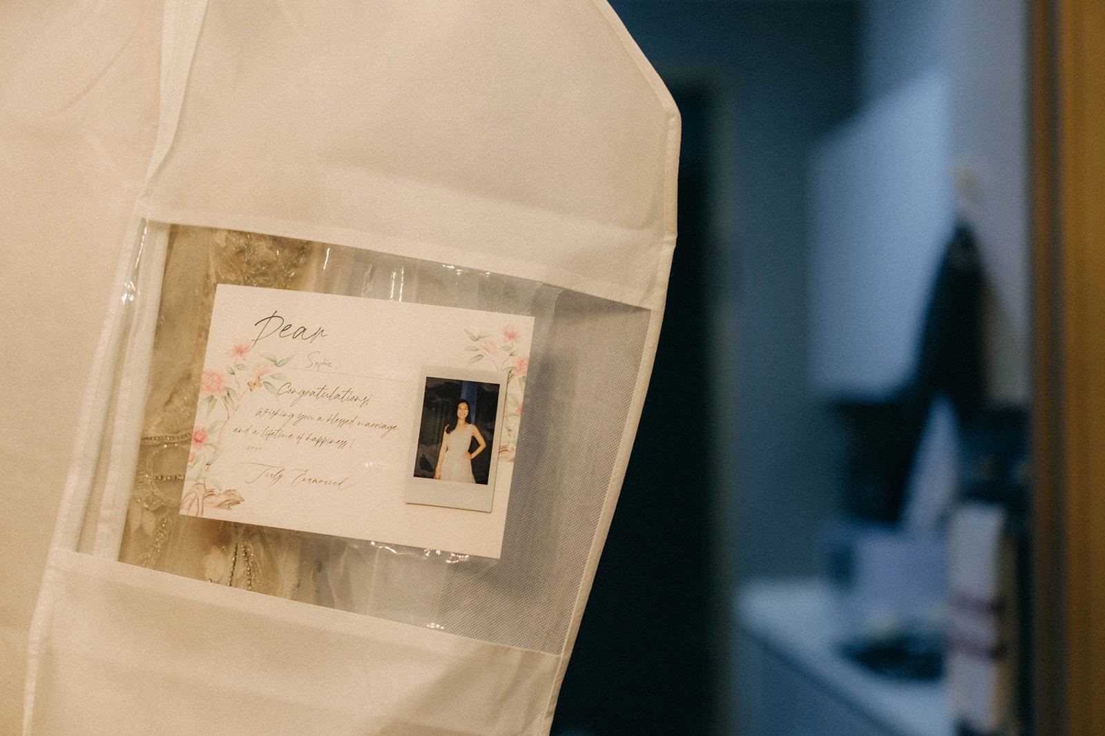 A wedding dress in a protective garment bag, with a handwritten card addressed to Sophie congratulating her on her marriage, featuring a polaroid photo of the bride. This scene captures a moment before a wedding at Monti at 1-Pavilion, Singapore.