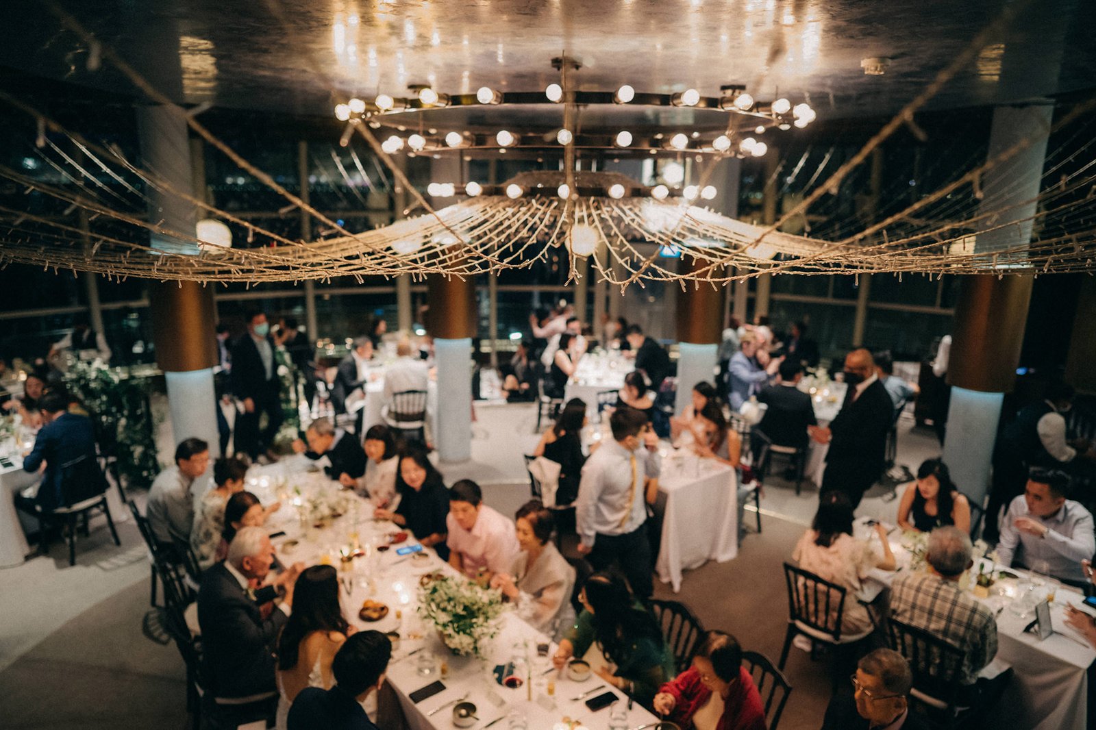 Elegant wedding reception at Monti at 1-Pavilion, Singapore, with guests seated at round tables adorned with floral centerpieces, under a grand chandelier.