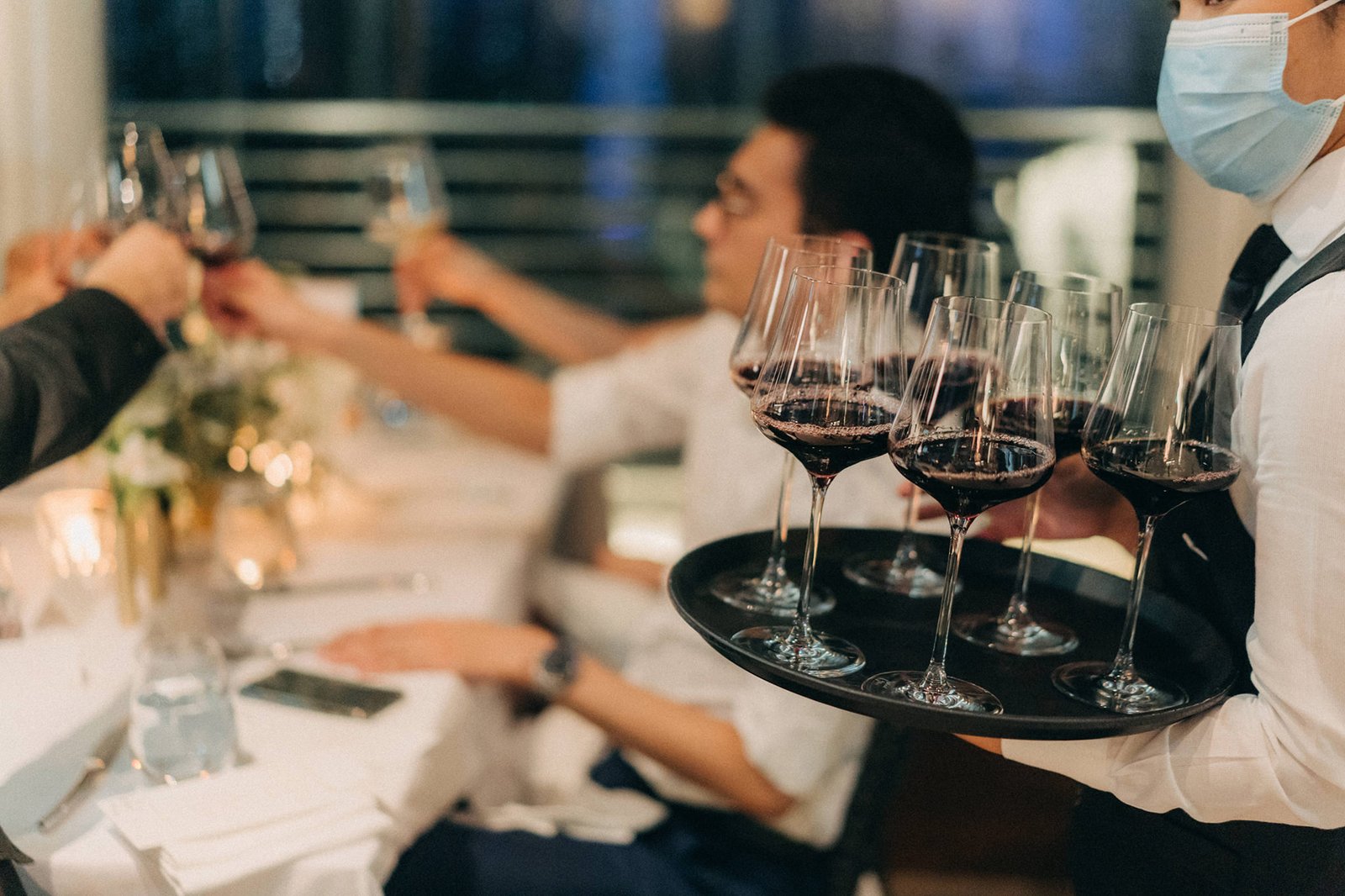 A server in a mask offers a tray of red wine glasses at a Monti at 1-Pavilion wedding in Singapore, with guests toasting in the background.