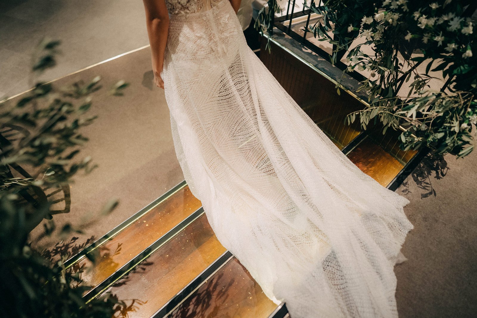 Bride in a detailed lace wedding gown with a long train walks down illuminated stairs at a Monti at 1-Pavilion wedding in Singapore. Lush greenery frames the scene.