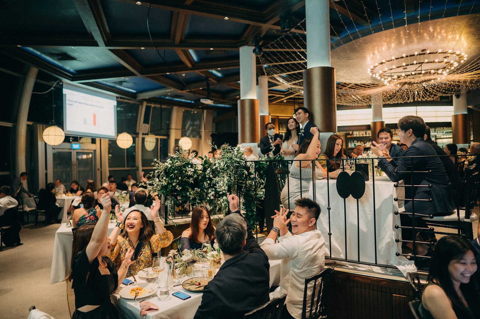 Wedding guests cheer and laugh at a lively reception dinner at Monti, a popular wedding venue in Singapore. A bride in a white dress and groom in a suit are visible in the background.