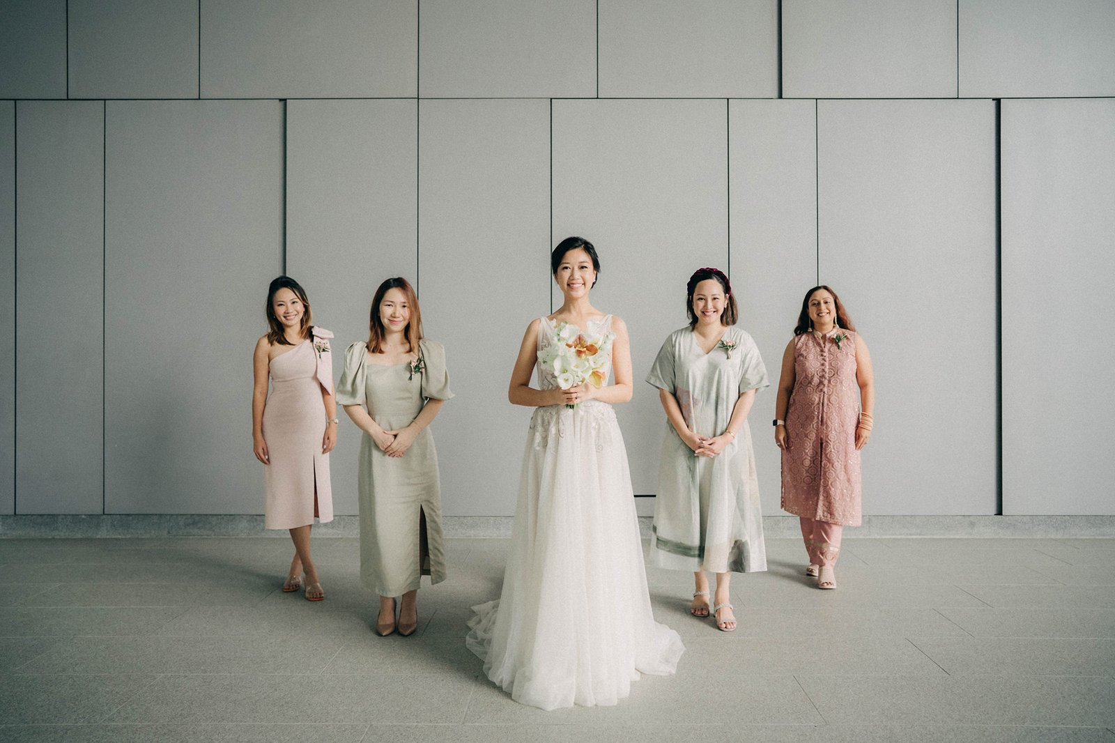 Bride in a white wedding dress holds a bouquet, flanked by five women in varying pastel dresses, posing against a modern, gray wall at a Monti at 1-Pavilion wedding in Singapore.