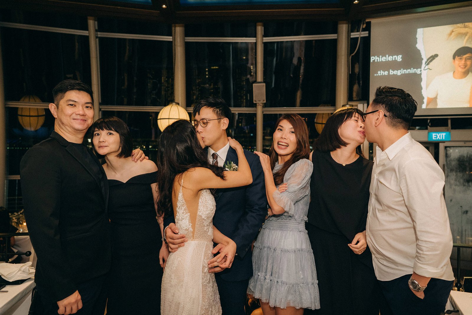 A wedding party poses for a photo at Monti at 1-Pavilion, Singapore. The bride and groom share a kiss in the center, surrounded by their friends and family. A projection of the couple is visible in the background.