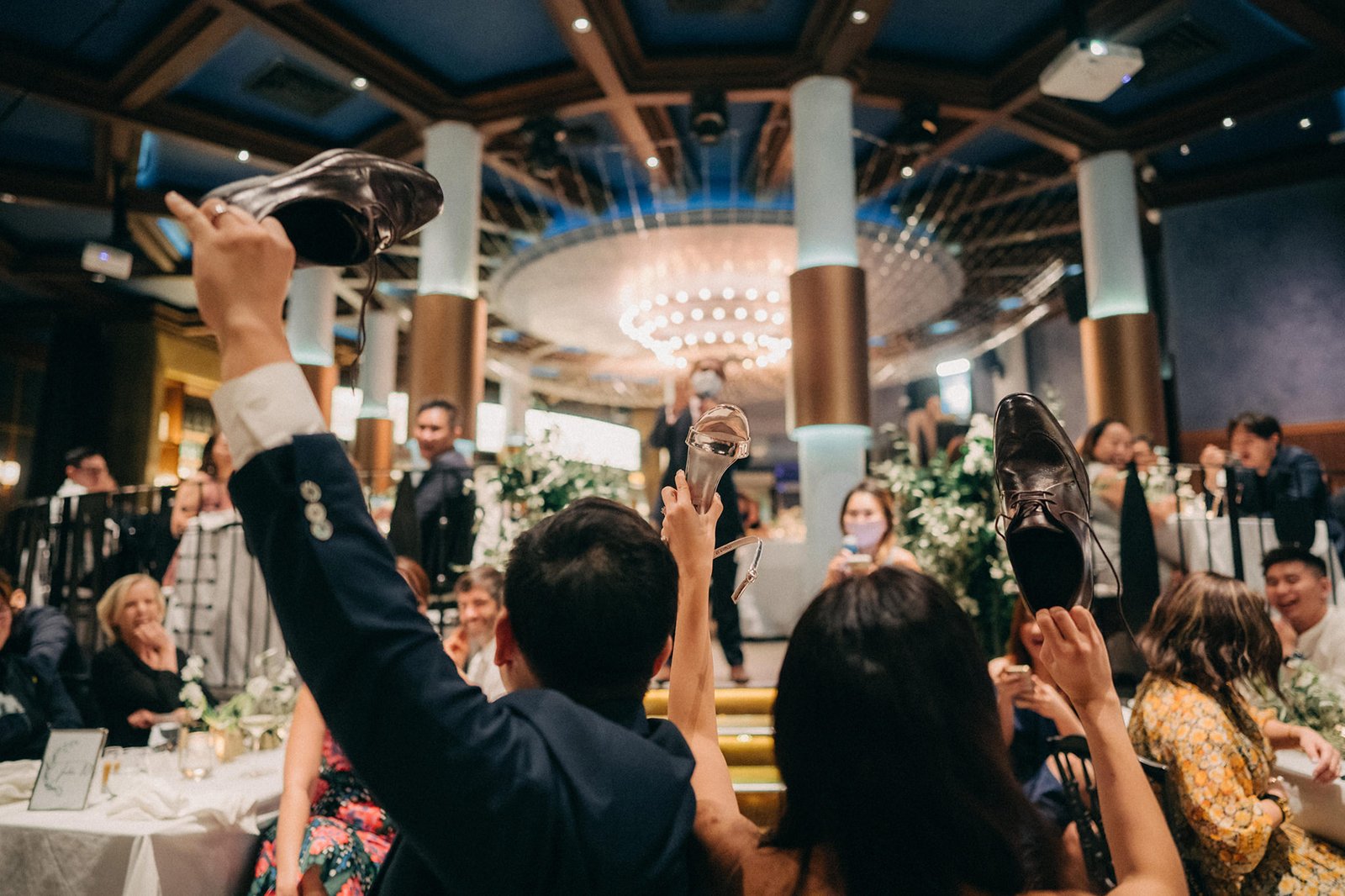 Wedding guests at Monti at 1-Pavilion, Singapore, playfully raise shoes during a reception, celebrating LK and Sophie's special day. The elegant venue features a grand chandelier and pillar decor.