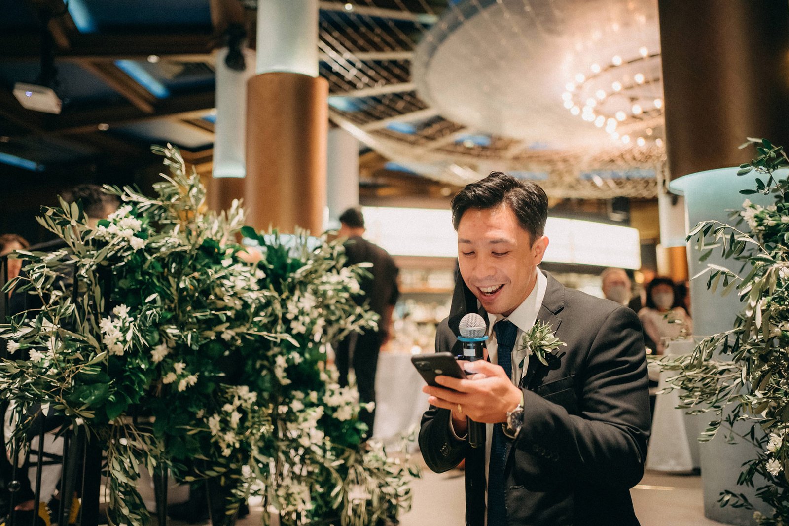 A smiling groom in a suit holds a phone and speaks into a microphone at his wedding reception at Monti at 1-Pavilion, Singapore, surrounded by lush greenery.
