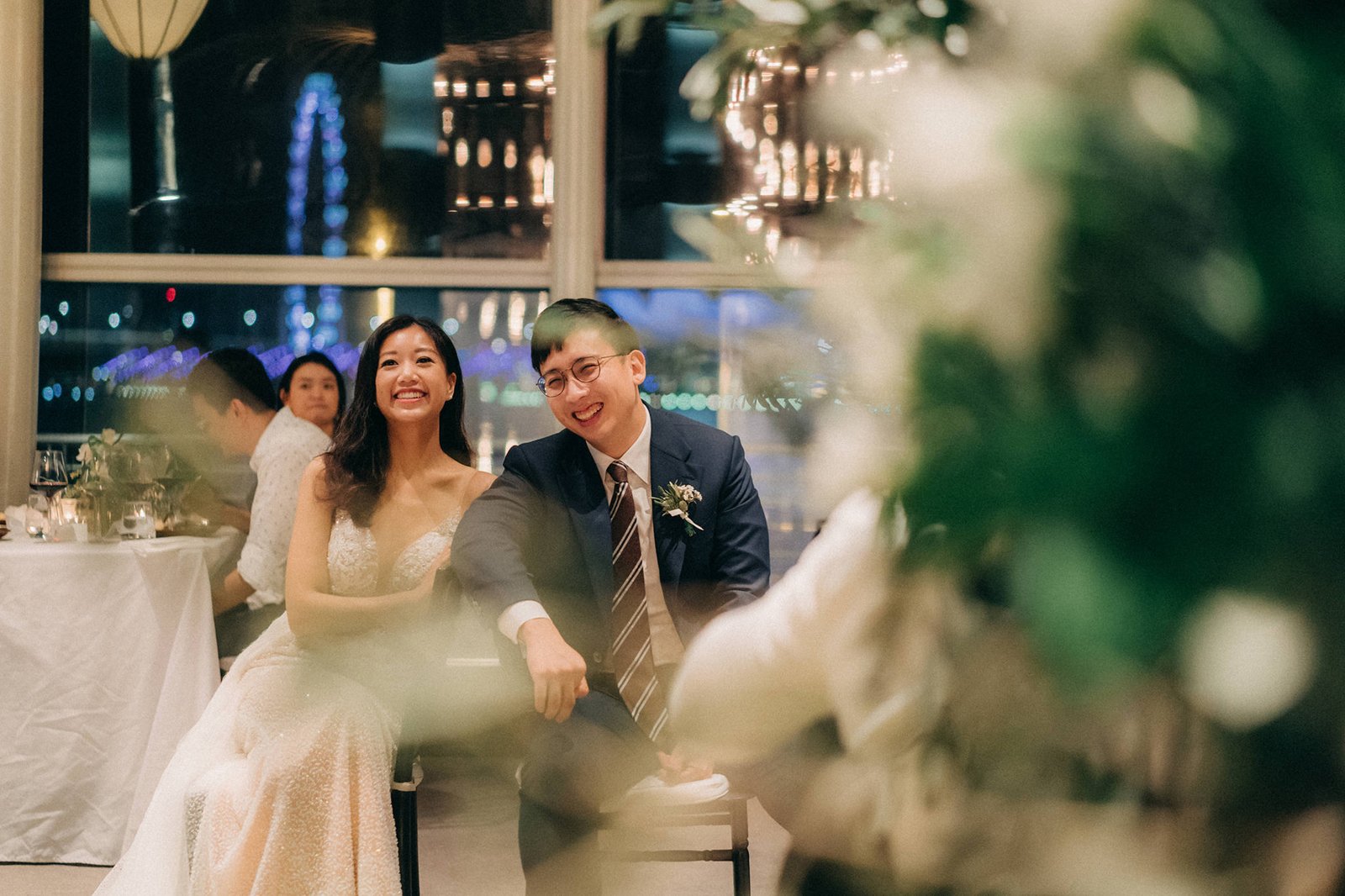 A smiling couple, LK and Sophie, at their Monti wedding in Singapore. Sophie wears a sparkling gown, and LK is in a suit, both looking towards guests at their reception. The background shows city lights and elegant decor.