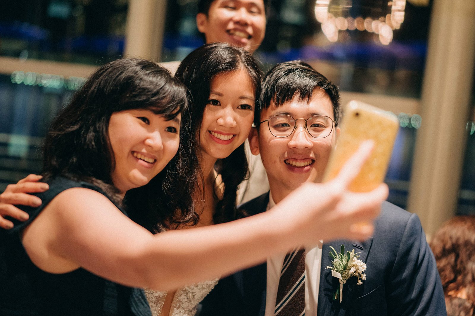 A happy group of three friends take a selfie at a wedding reception at Monti, Singapore, with the city skyline blurred in the background. The bride, Sophie, beams between two friends at their Monti at 1-Pavilion wedding celebration.