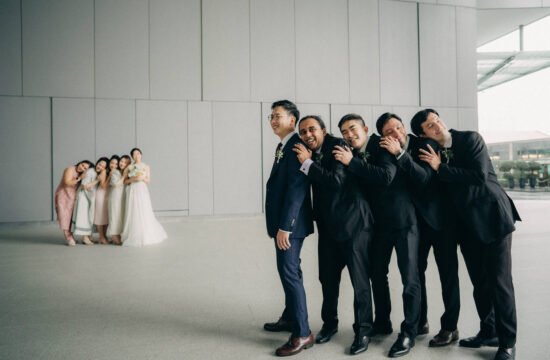 Groomsmen in black suits playfully lean against each other in line, facing a wedding party in soft pastel dresses. Monti at 1-Pavilion wedding Singapore photography.