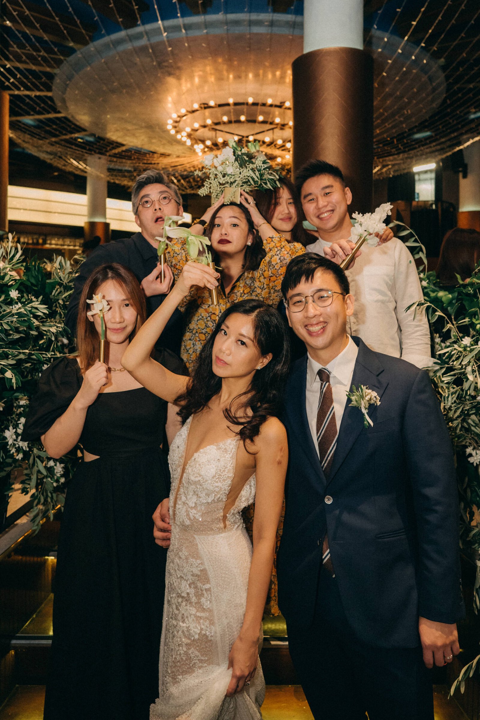 Wedding party at Monti at 1-Pavilion, Singapore, with the bride in a lace gown and groom in a navy suit, surrounded by friends holding flowers.