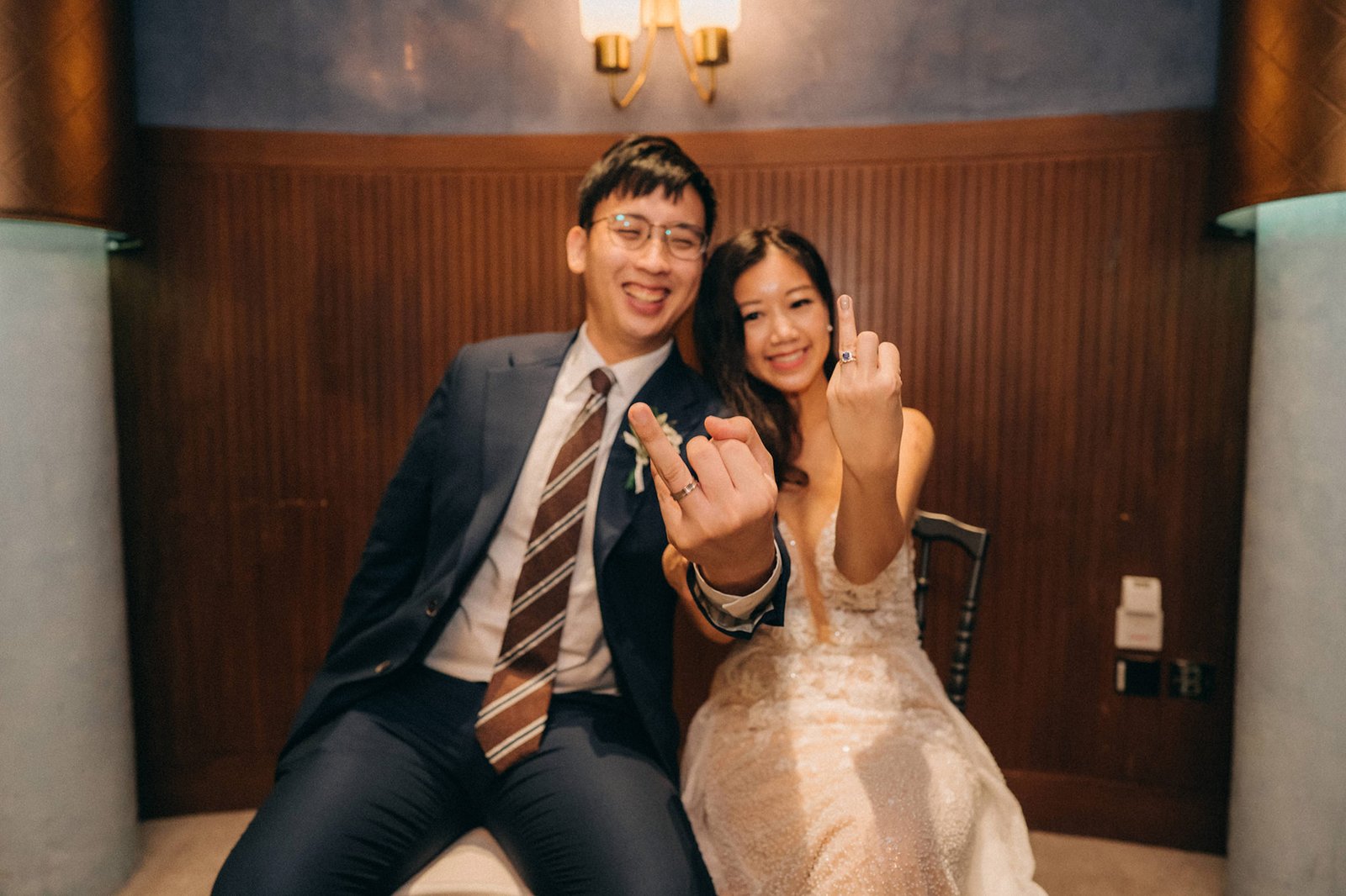 A joyful bride and groom at their Monti at 1-Pavilion wedding in Singapore, showing off their wedding rings and giving the middle finger with a playful expression. The groom wears a navy suit and striped tie, while the bride is in a sparkling white wedding dress.