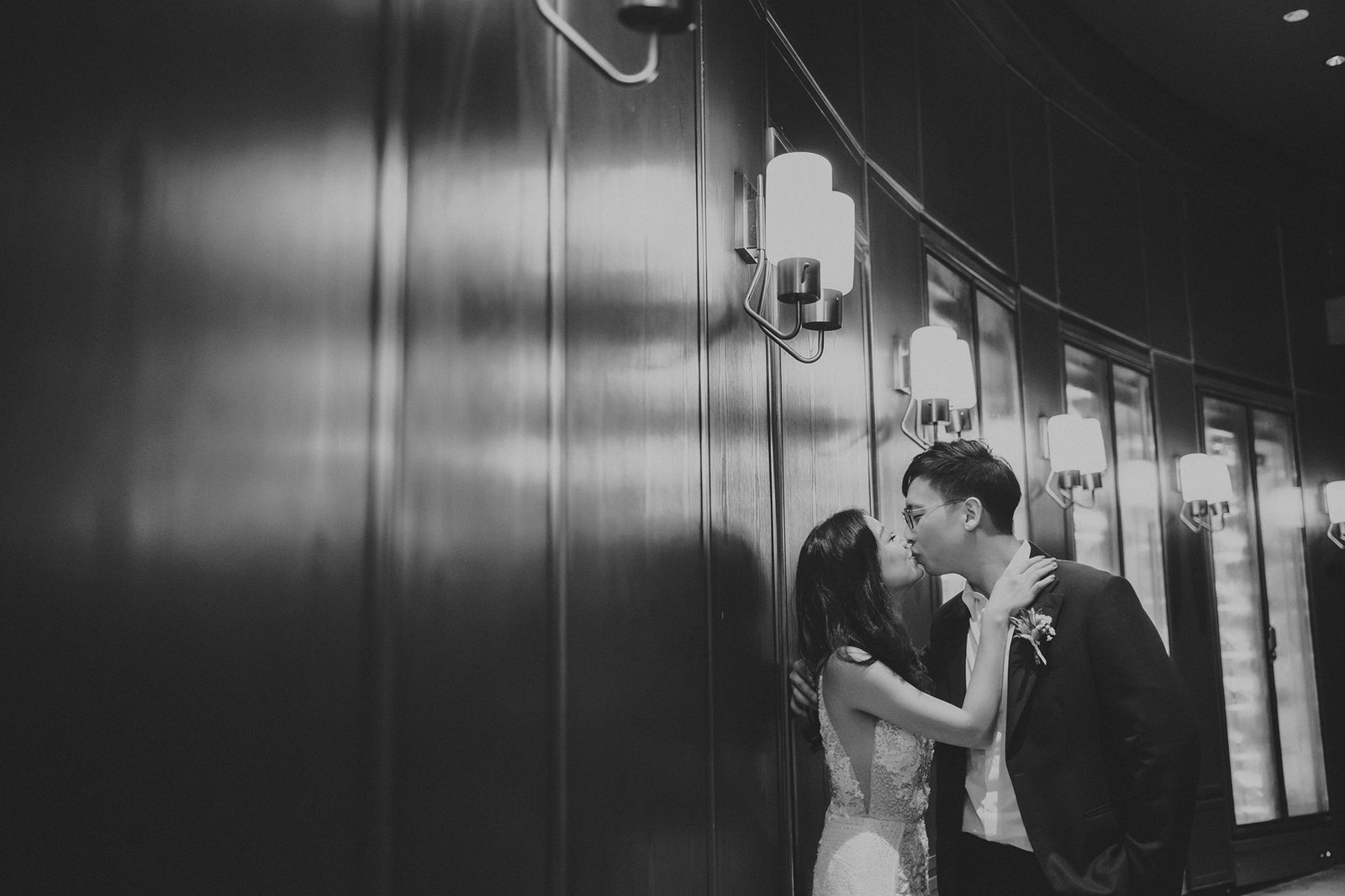 Black and white wedding photography at Monti at 1-Pavilion, Singapore: A bride in a lace gown and groom share a tender kiss against a dark, wood-paneled wall with elegant sconces.