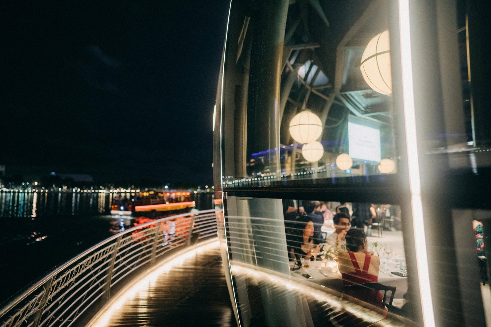 Elegant wedding reception at Monti at 1-Pavilion, Singapore, with guests dining indoors reflected in a large glass window overlooking a nighttime cityscape and waterfront. Monti at 1-Pavilion wedding photography.