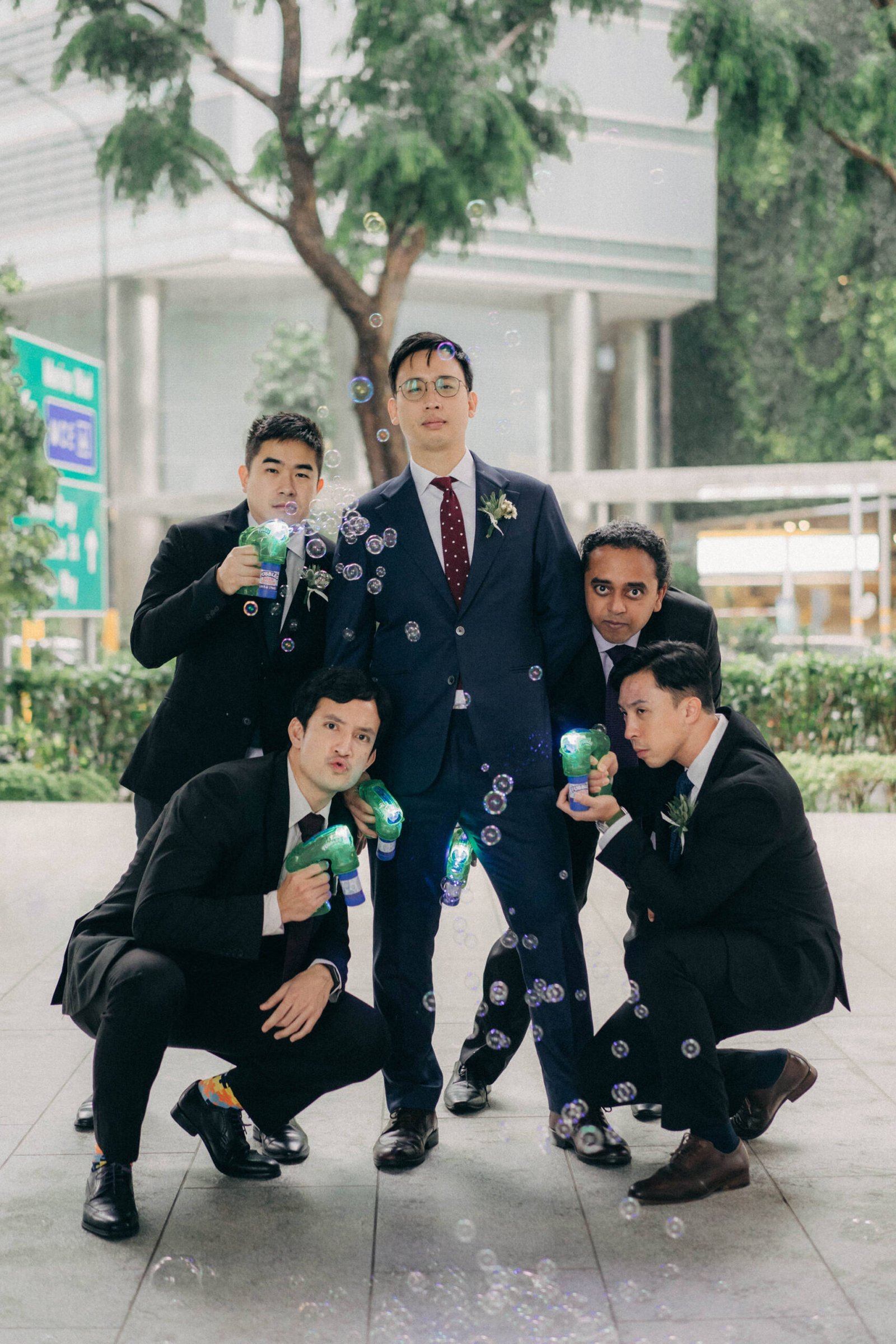 Groomsmen at a Monti at 1-Pavilion wedding in Singapore, holding bubble guns and having fun. The groom stands center in a navy suit, surrounded by his smiling groomsmen in black suits.