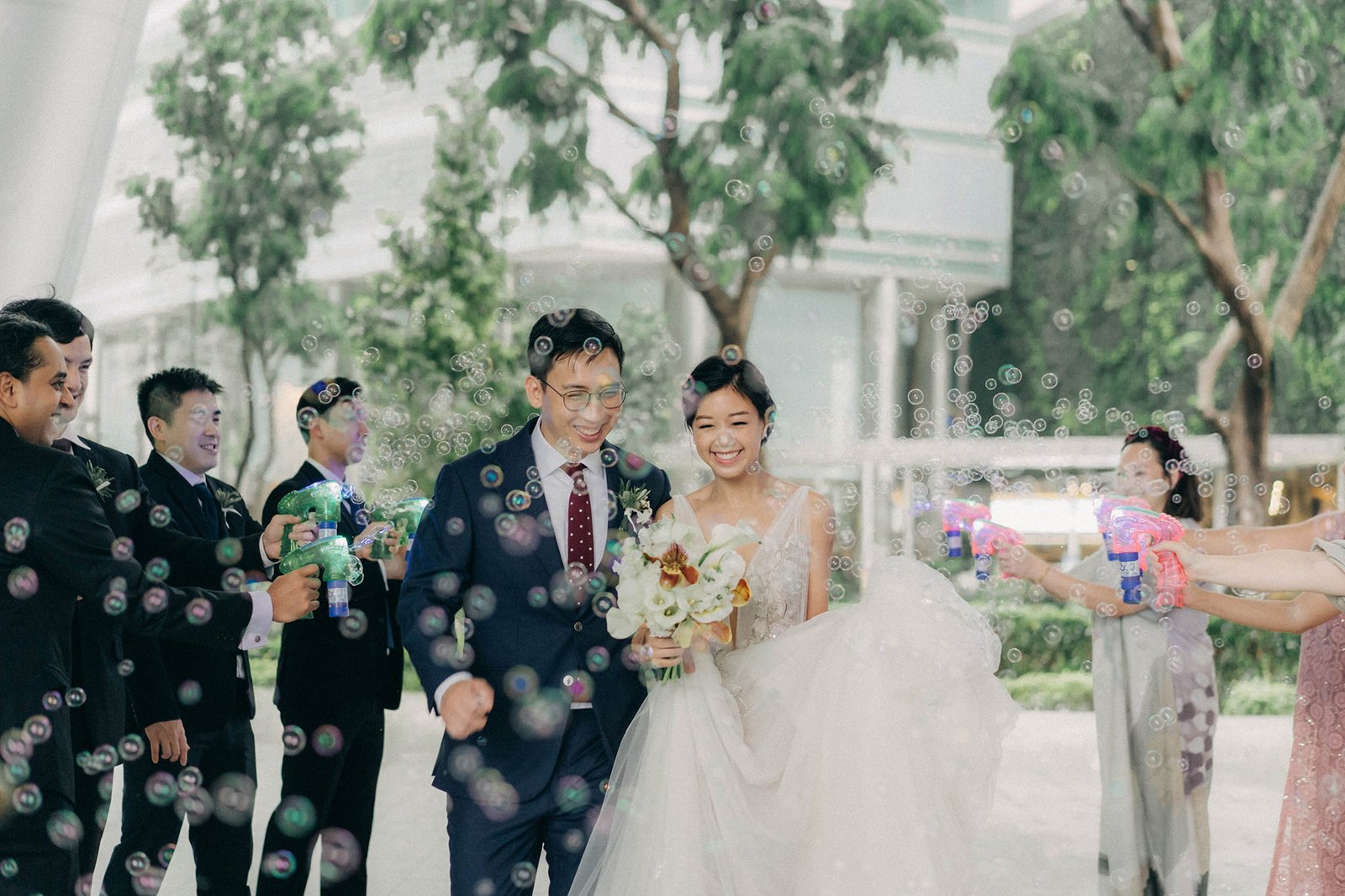 A smiling bride and groom walk through a shower of bubbles at their Monti at 1-Pavilion wedding in Singapore, captured by photography.