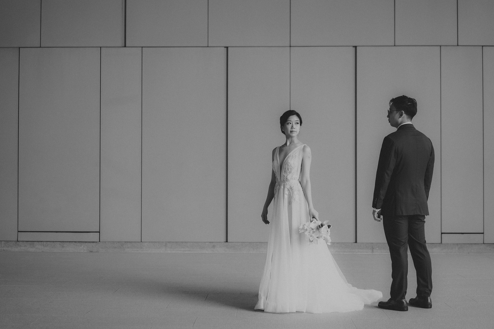 Black and white wedding photography at Monti at 1-Pavilion, Singapore, shows a bride in a lace gown holding a bouquet, looking away from a groom in a suit.
