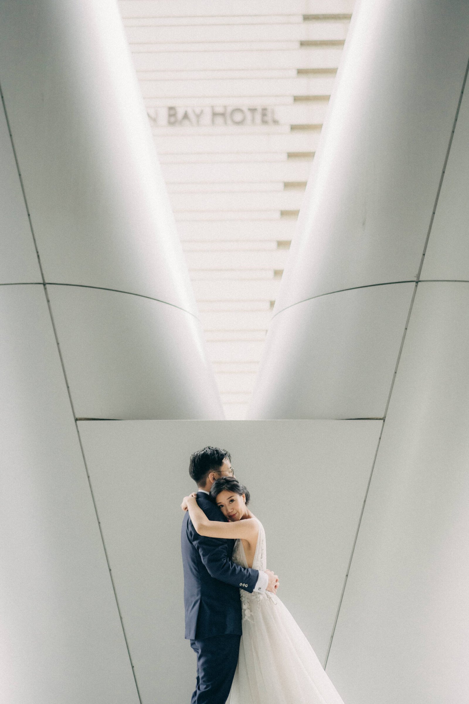 Bride and groom embracing in front of the Monti at 1-Pavilion, Singapore, during their wedding photography session. The bride wears a white gown, and the groom a navy suit.