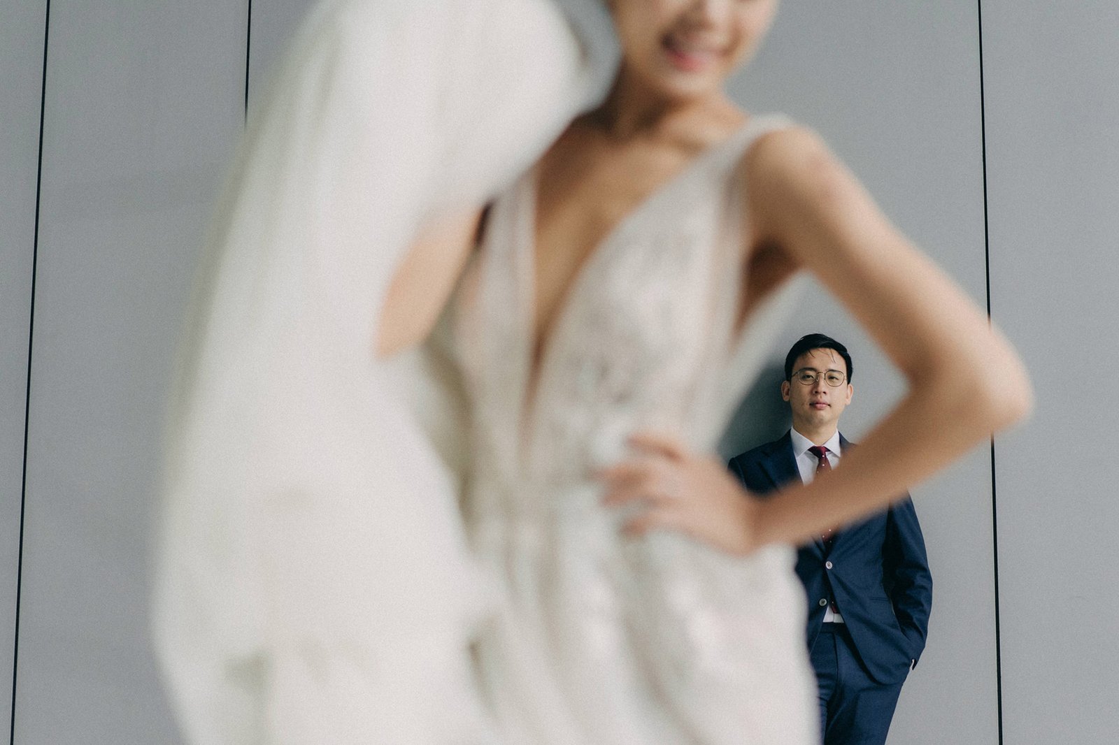 A bride in a flowing white gown is in the foreground, out of focus, with her groom in a navy suit and red tie standing behind her and smiling. This is a wedding photography scene at Monti at 1-Pavilion, Singapore.