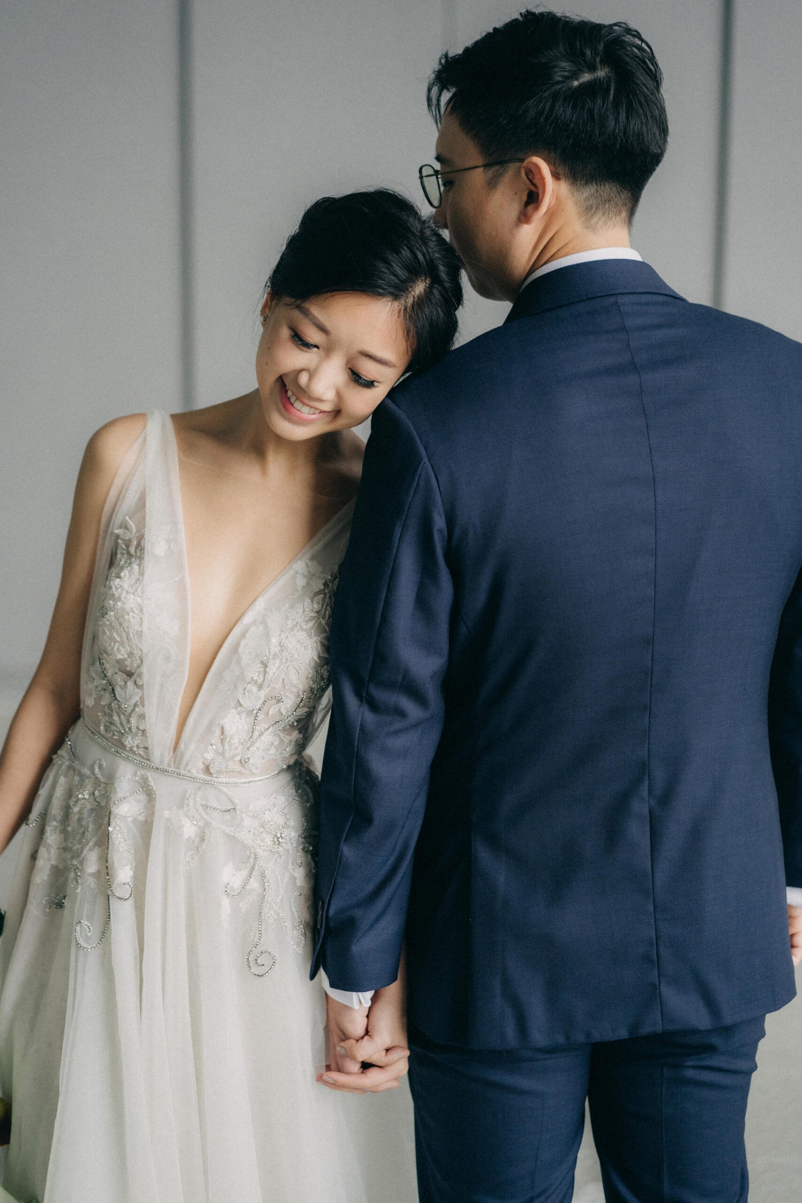 A joyful bride in a detailed white wedding gown rests her head on the shoulder of a groom in a navy suit, holding hands at their Monti at 1-Pavilion wedding in Singapore, captured by wedding photography.