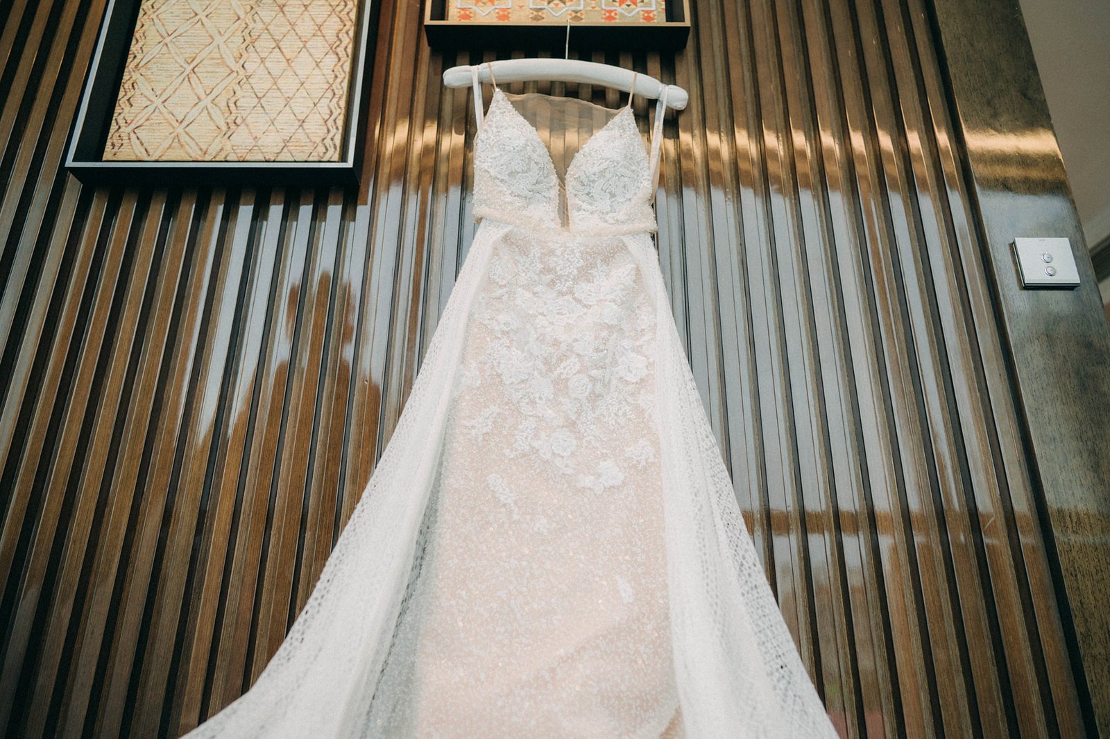 Elegant white lace wedding dress with intricate beading hangs on a hanger against a textured wooden wall at Monti at 1-Pavilion, Singapore, a beautiful wedding venue.