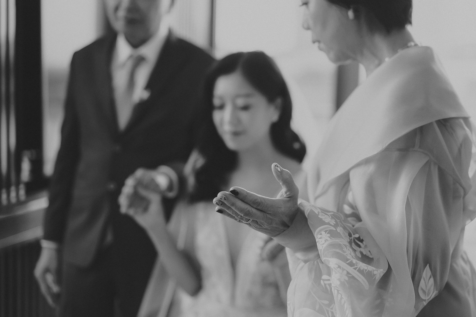 A bride, her mother, and father share a tender moment during a wedding ceremony at Monti at 1-Pavilion, Singapore. The bride's mother gently holds her hand, her fingers splayed, as the bride looks down. The father stands beside them, his arm around the bride. The scene captures the emotional essence of a wedding celebration.