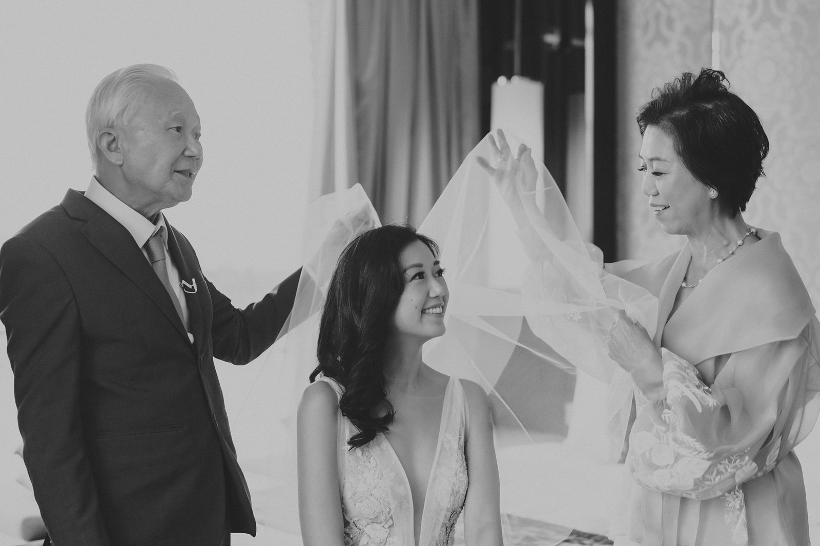 A bride smiles joyfully as her parents help her with her wedding veil during a Monti at 1-Pavilion wedding in Singapore. The black and white photo captures a tender moment before the ceremony.
