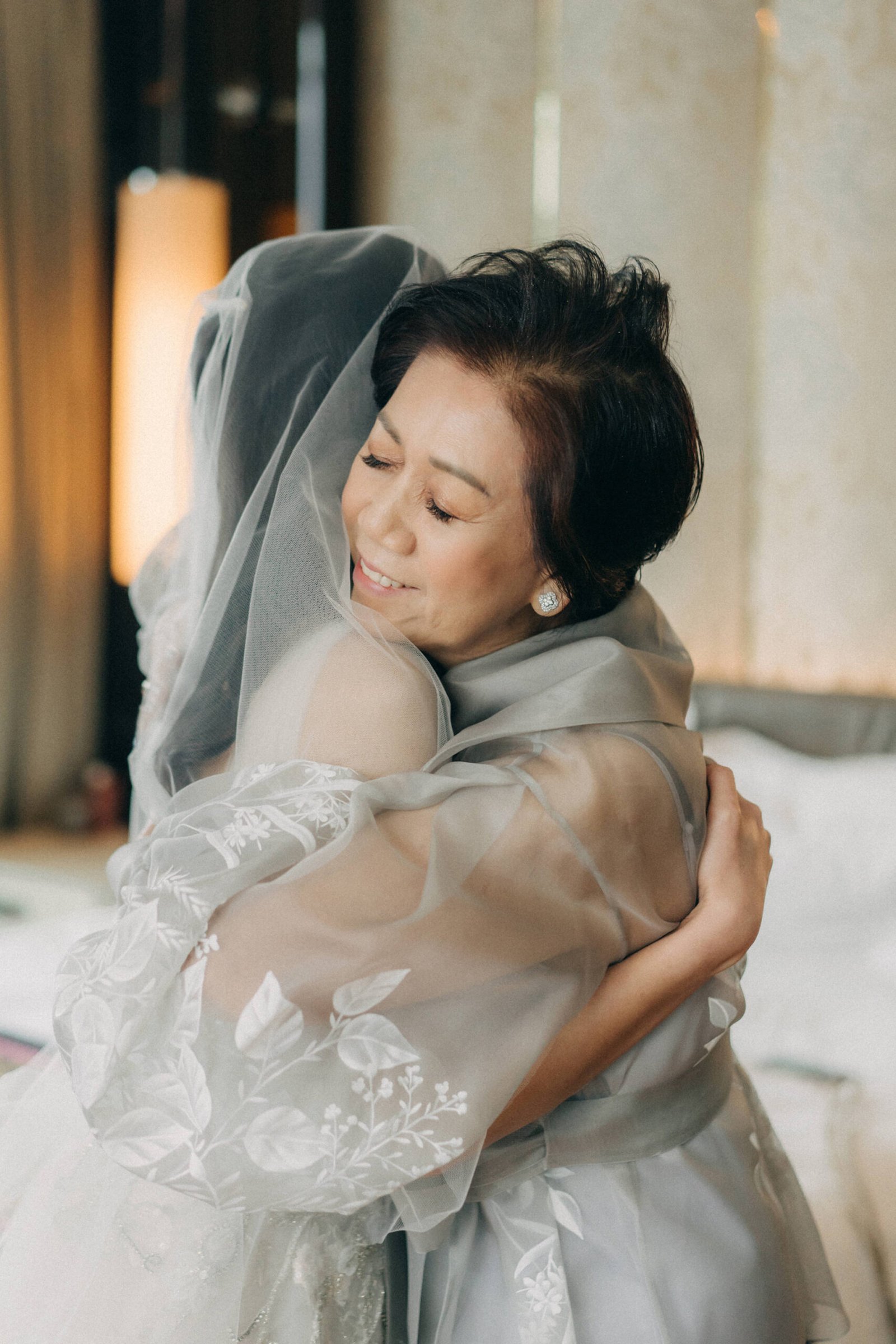 A mother in a grey dress tenderly hugs her daughter, the bride in a white gown and veil, during a Monti Circles wedding in Singapore. Photography by Monti at 1-Pavilion.