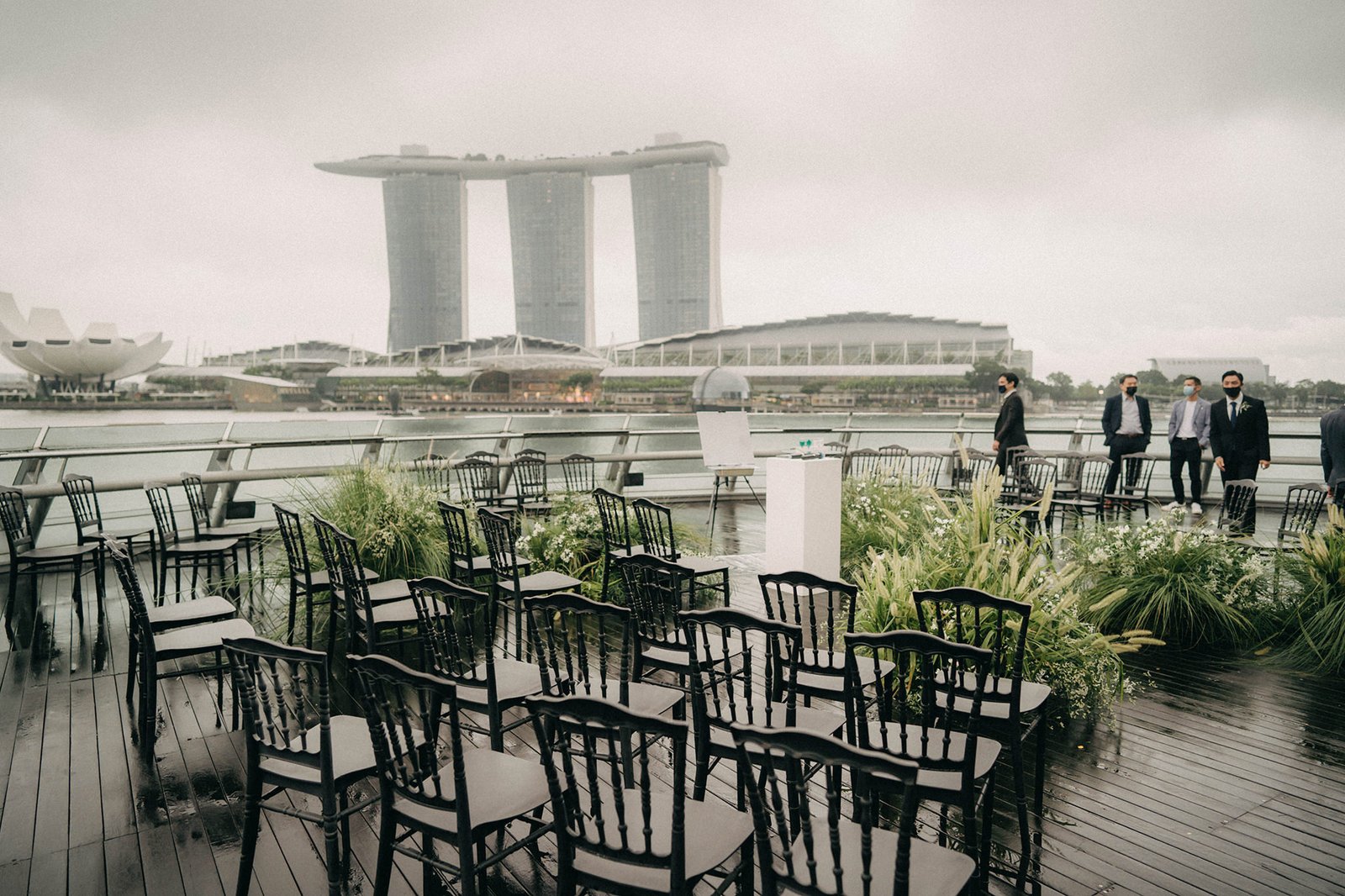 Outdoor wedding ceremony setup at Monti at 1-Pavilion, Singapore, with chairs arranged on a wet wooden deck, lush greenery, and the iconic Marina Bay Sands in the background. Wedding photography by [Photographer's Name].