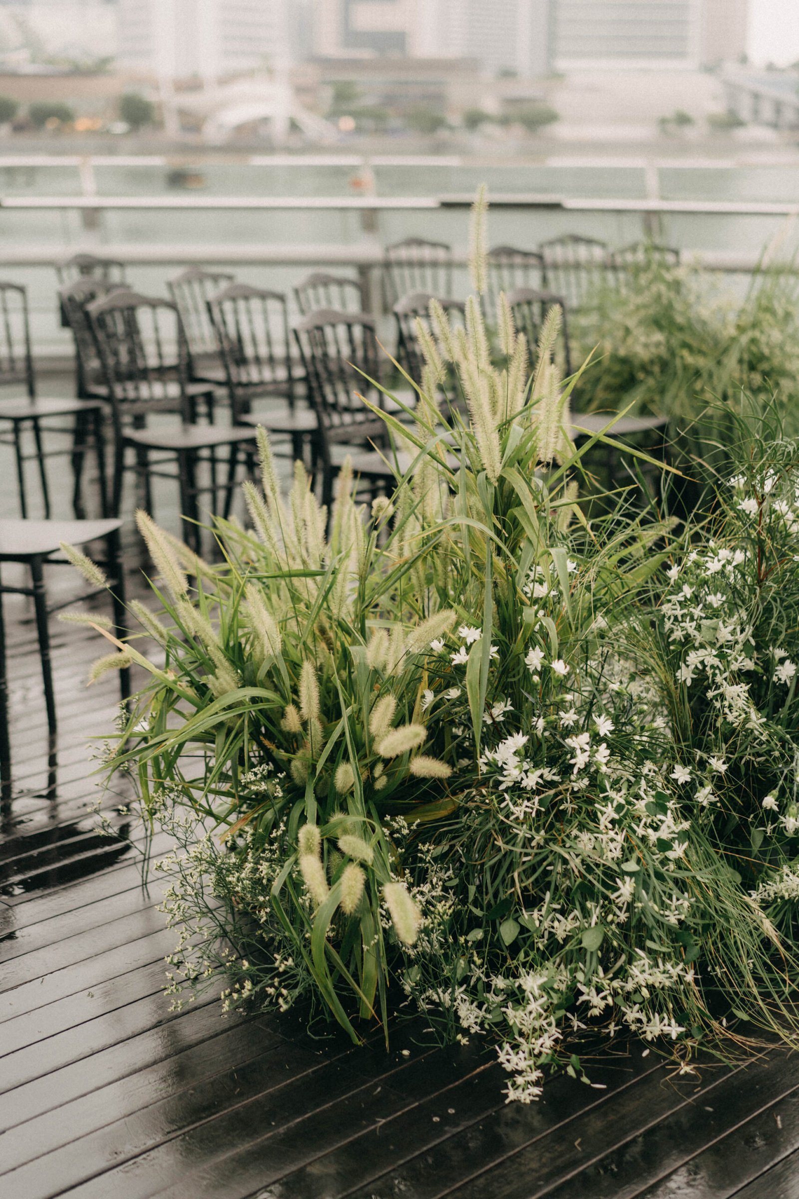 Outdoor wedding ceremony setup at Monti at 1-Pavilion, Singapore, featuring lush green floral arrangements and rows of chairs on a wet wooden deck.